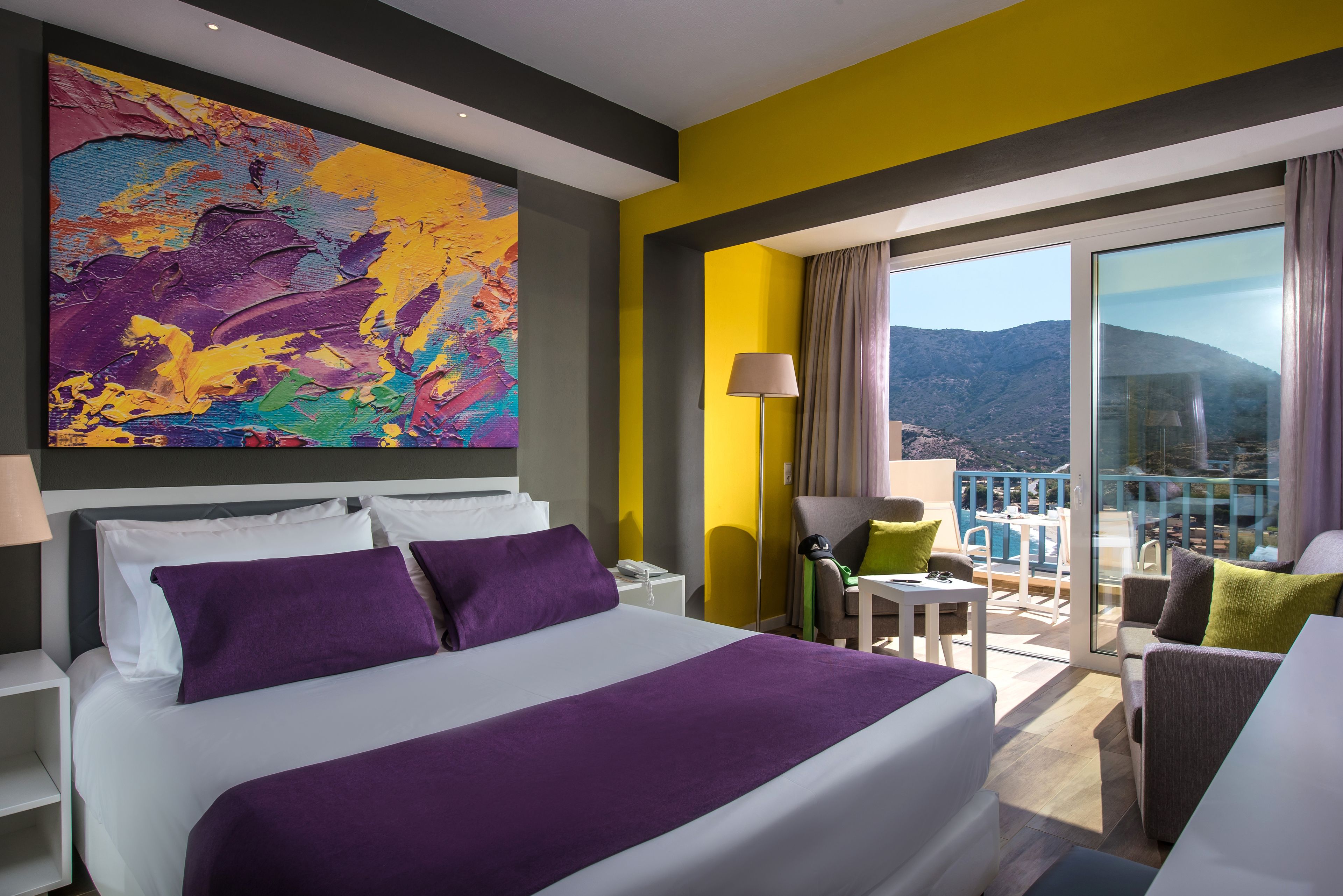 A hotel room with a bed, a colorful abstract painting, a balcony with a view of mountains, and a chair.