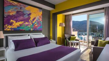 A hotel room with a bed, a colorful abstract painting, a balcony with a view of mountains, and a chair.