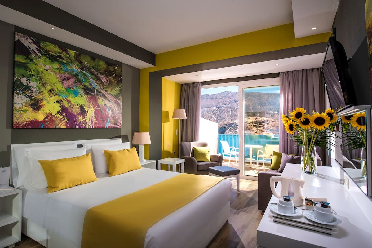 A modern hotel room with a large bed, a balcony with a view, and a dining area with a table set for breakfast.