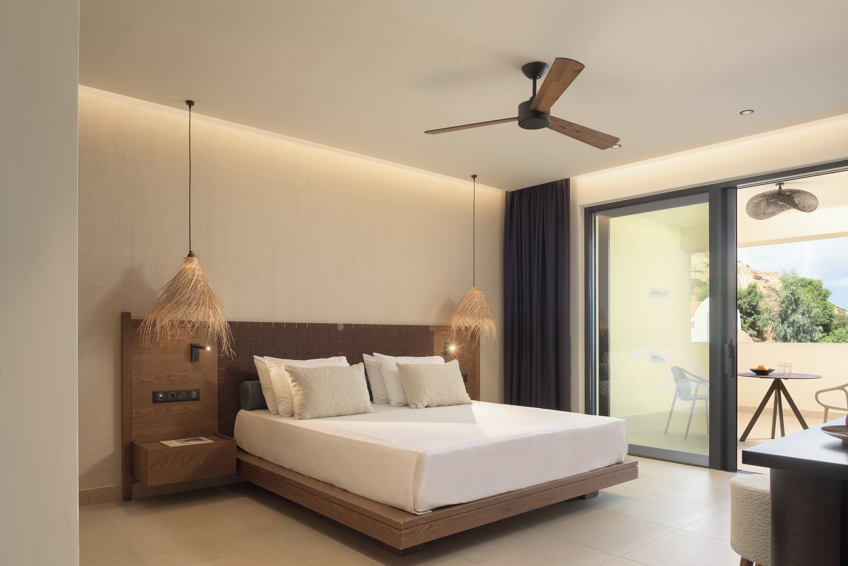 Une chambre d’hôtel moderne dotée d’un grand lit, d’un ventilateur de plafond et offrant une vue sur l’extérieur grâce à une porte-fenêtre coulissante.
