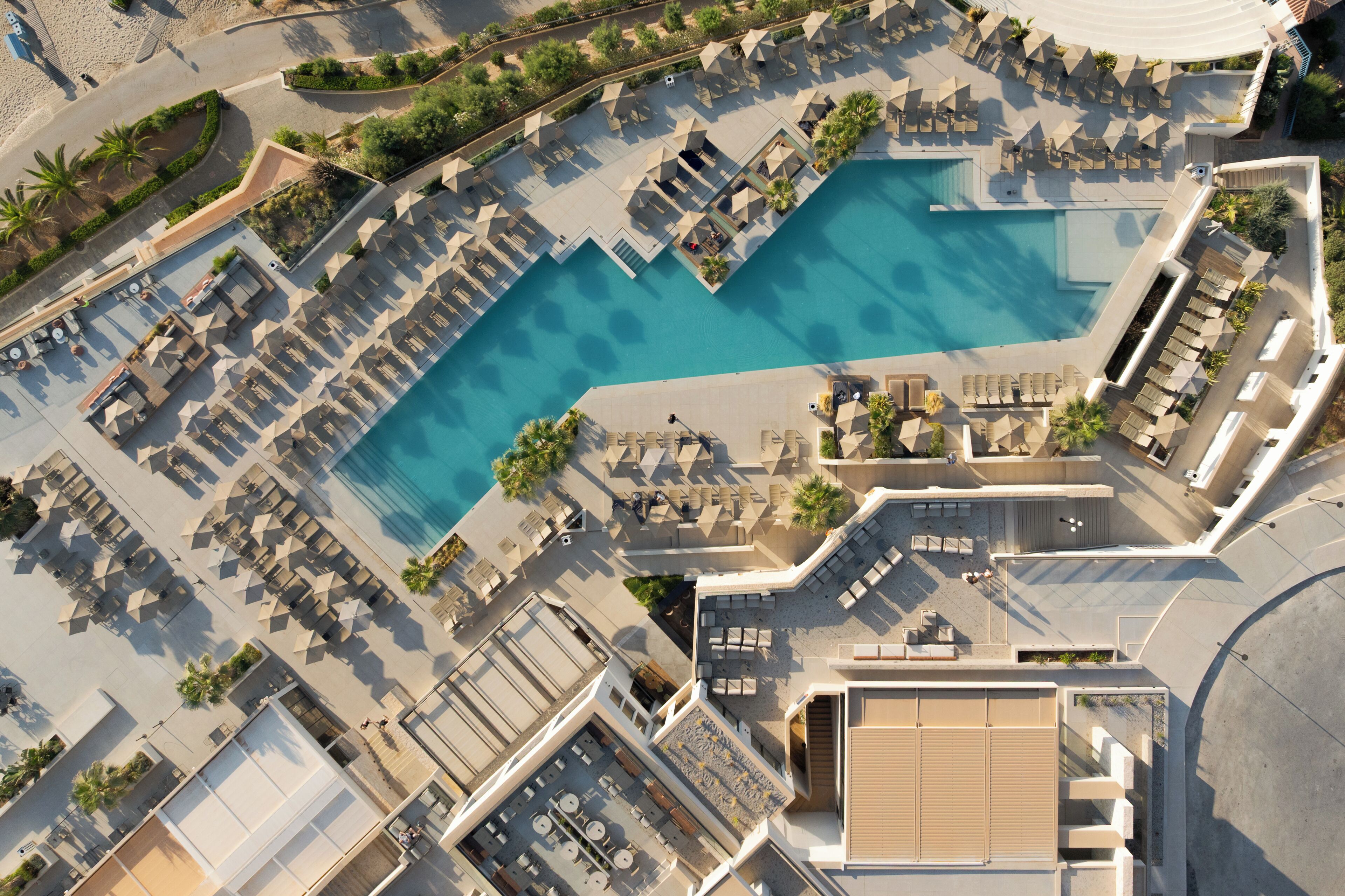 Vue aérienne d’un complexe hôtelier doté d’une grande piscine entourée de transats et de parasols, d’un espace de détente extérieur abrité et d’un jardin aménagé.