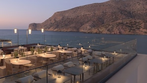 A rooftop terrace with tables and chairs, overlooking a body of water and a rocky cliff. - Fodele Beach & Water Park Holiday Resort - All Inclusive (Malevizi)
