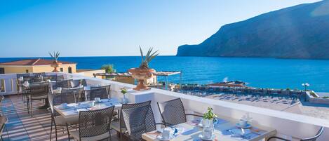 An outdoor terrace with tables and chairs, overlooking a body of water and a cliff.