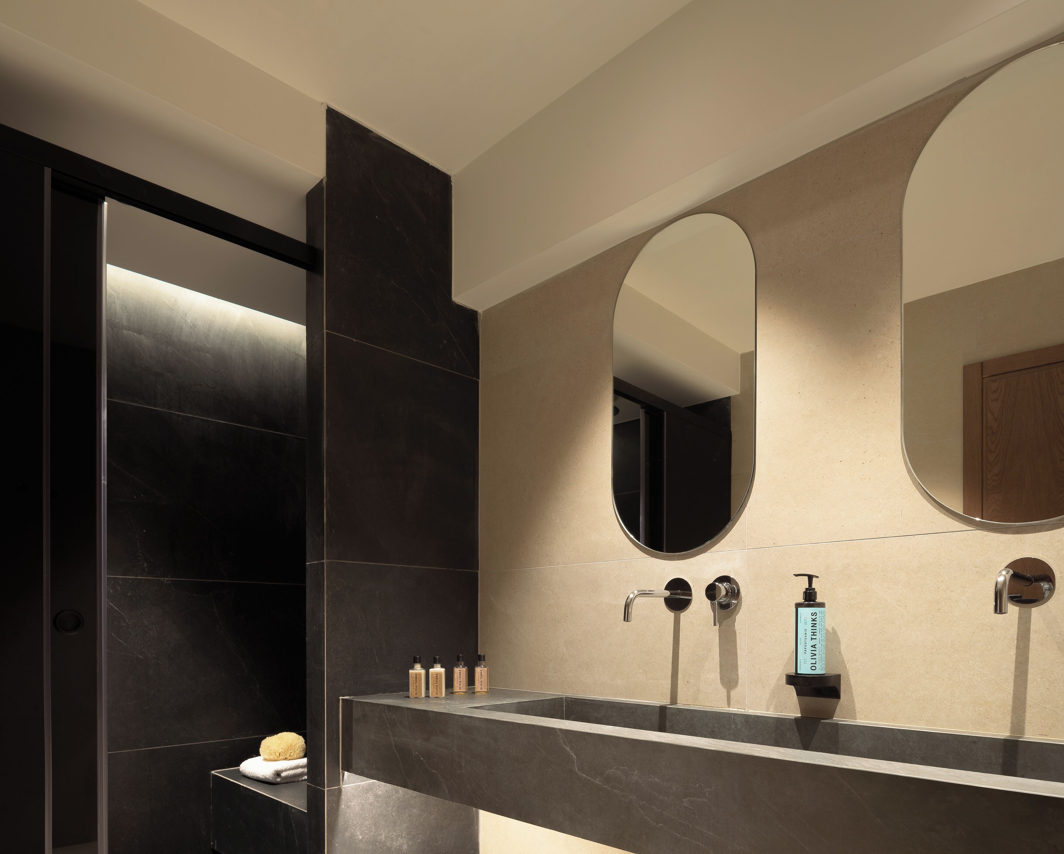 Une salle de bain moderne avec des carreaux foncés, un grand miroir et un lavabo doté de deux robinets et d’un distributeur de savon.