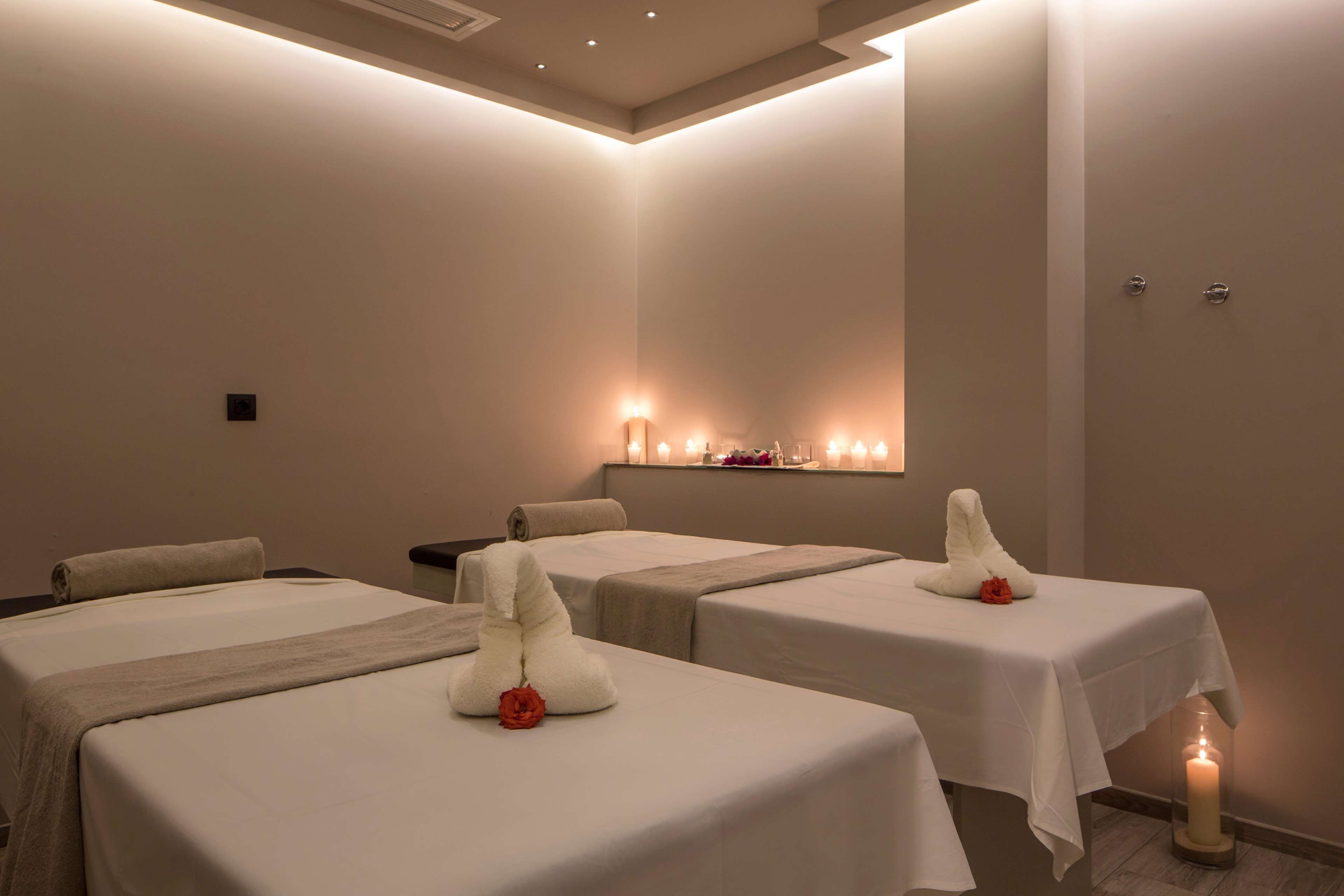 A spa room with three beds, each with a white towel and a red rose. There are candles on the floor and a shelf with candles in the background.