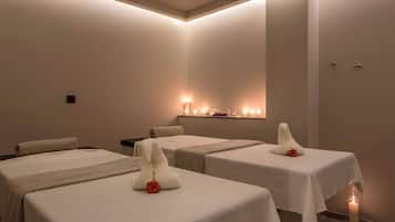A spa room with three beds, each with a white towel and a red rose. There are candles on the floor and a shelf with candles in the background.