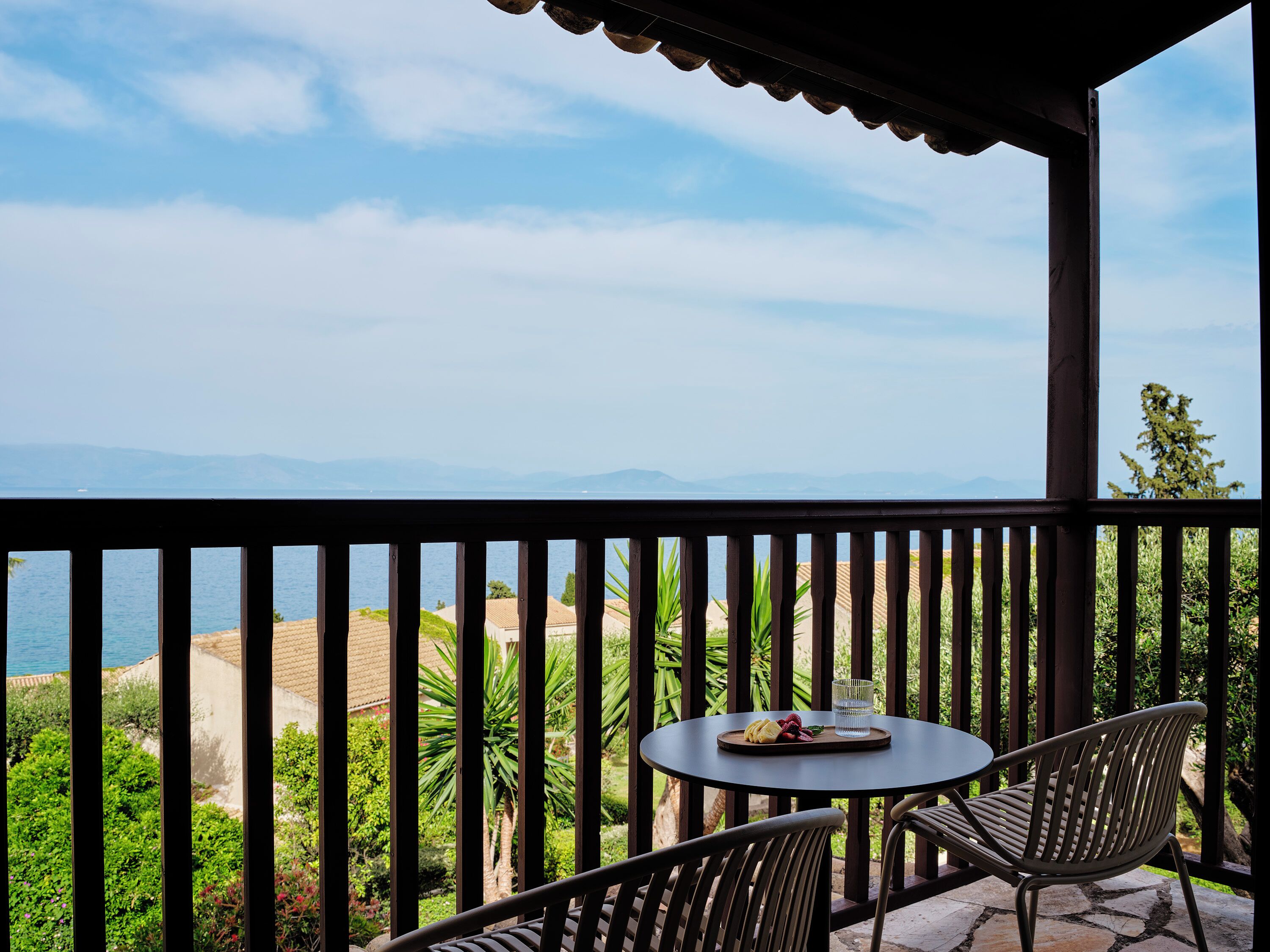 A balcony with a table and chairs, overlooking a coastal landscape with mountains in the distance.