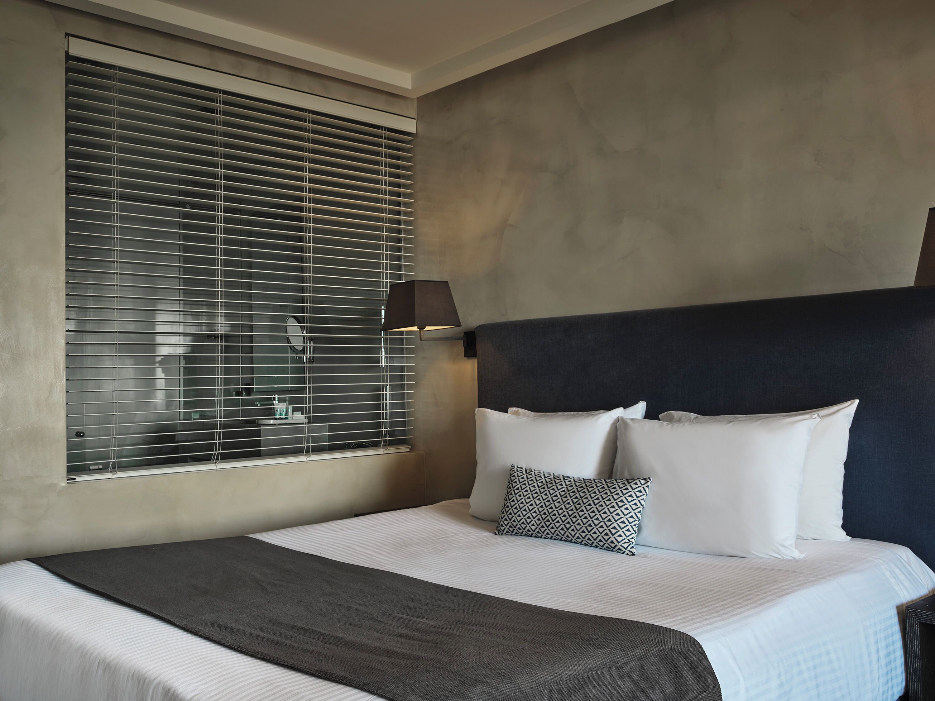 Habitación de hotel con una cama grande, cabecera, lámpara de noche y ventana con persianas.