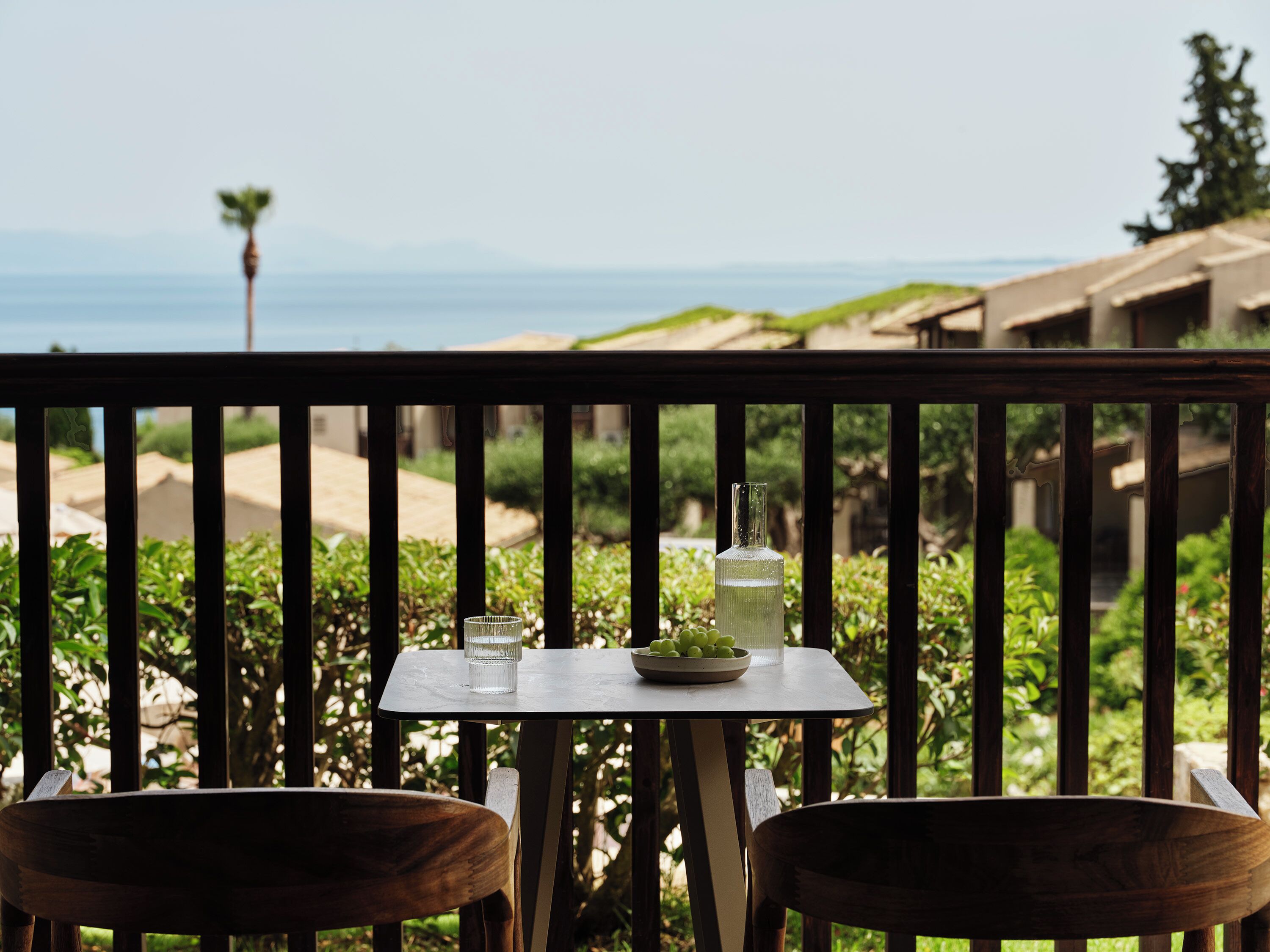 A balcony with a table and chairs, overlooking a landscaped garden and a distant view of the sea.