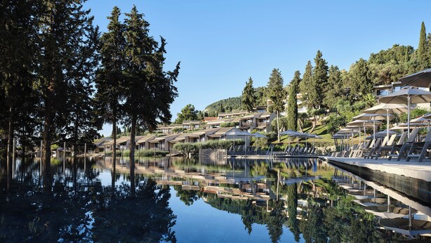 Un área de piscina tranquila con sillones de descanso y sombrillas, rodeada de árboles y una ladera con edificios.