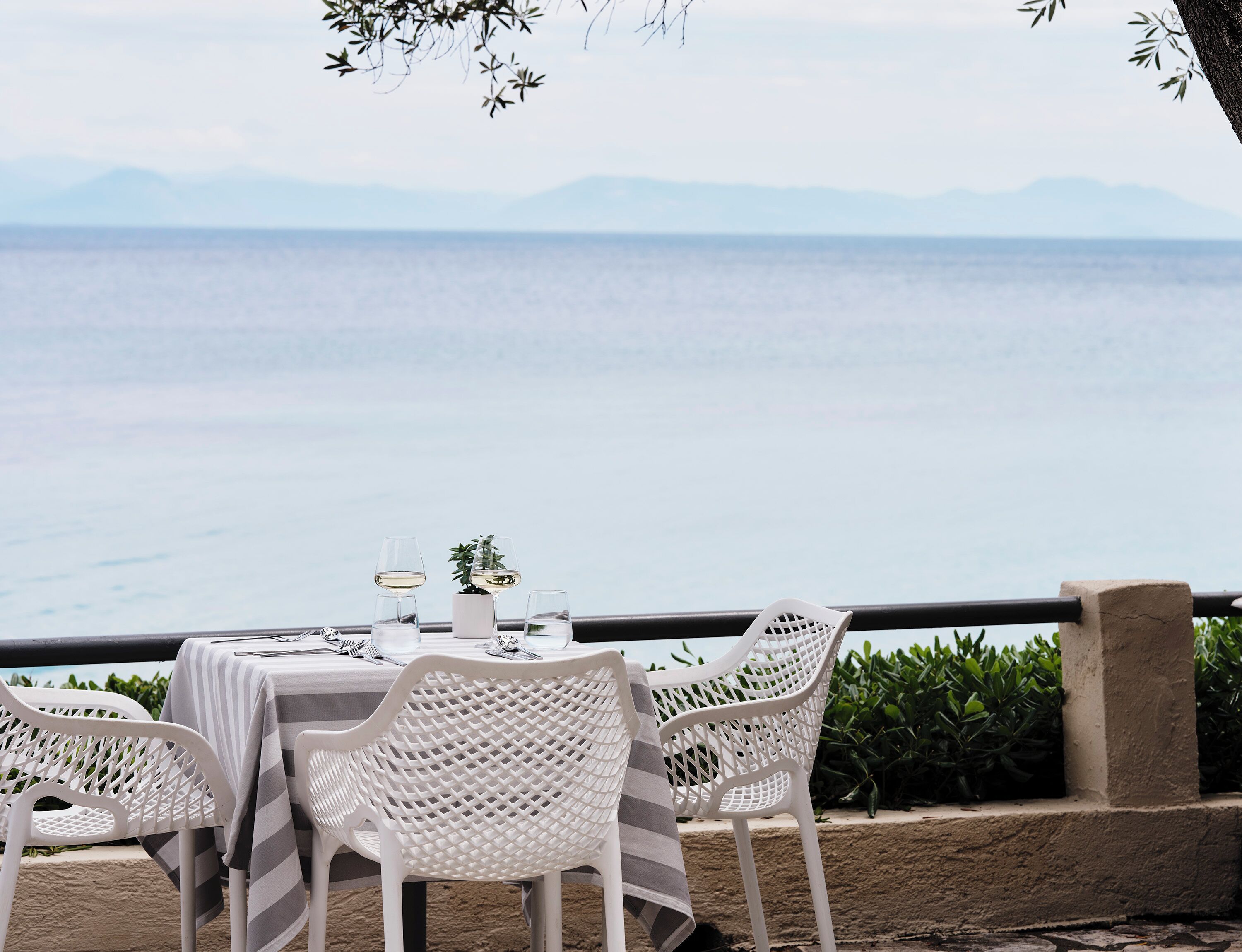 Une table dressée avec des chaises blanches et une nappe, donnant sur une étendue d’eau avec des montagnes à l’horizon.