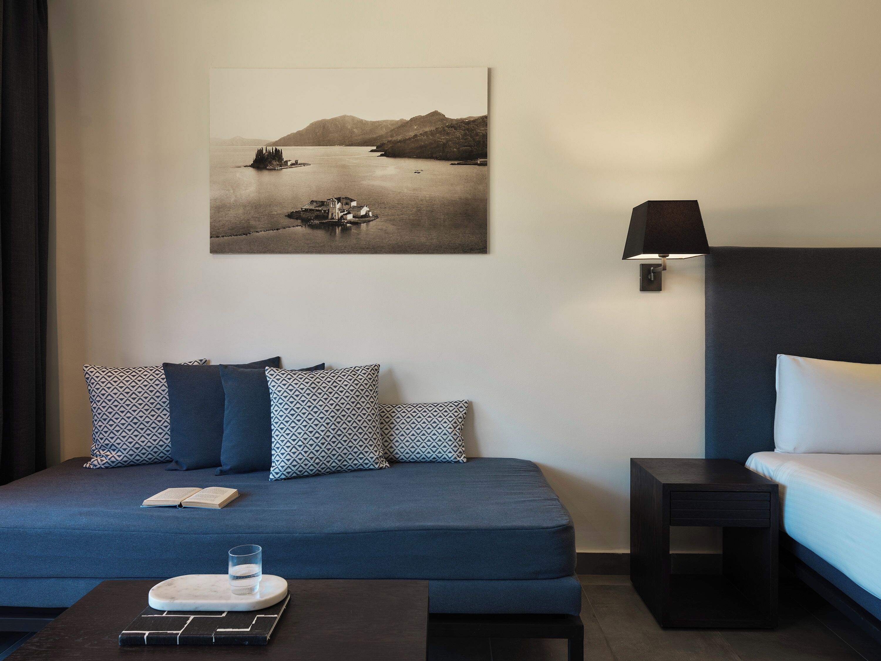 A modern bedroom with a blue sofa, patterned pillows, a bedside table, a lamp, and a framed landscape photograph on the wall.