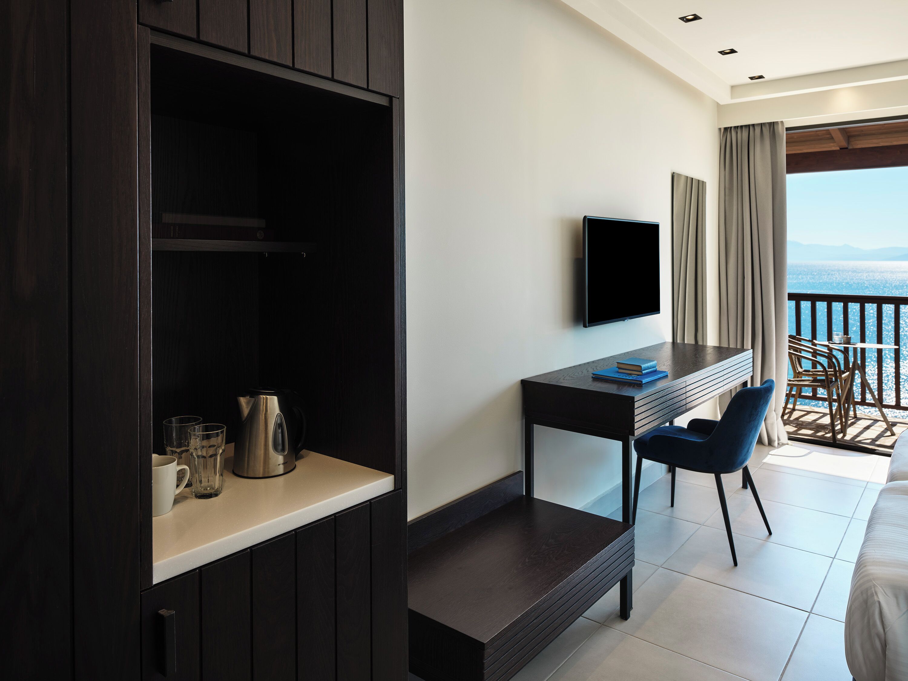 Habitación de hotel moderna con un escritorio de madera oscura, televisor de pantalla plana, una mesita con un libro, una silla azul y un balcón con vistas al mar.