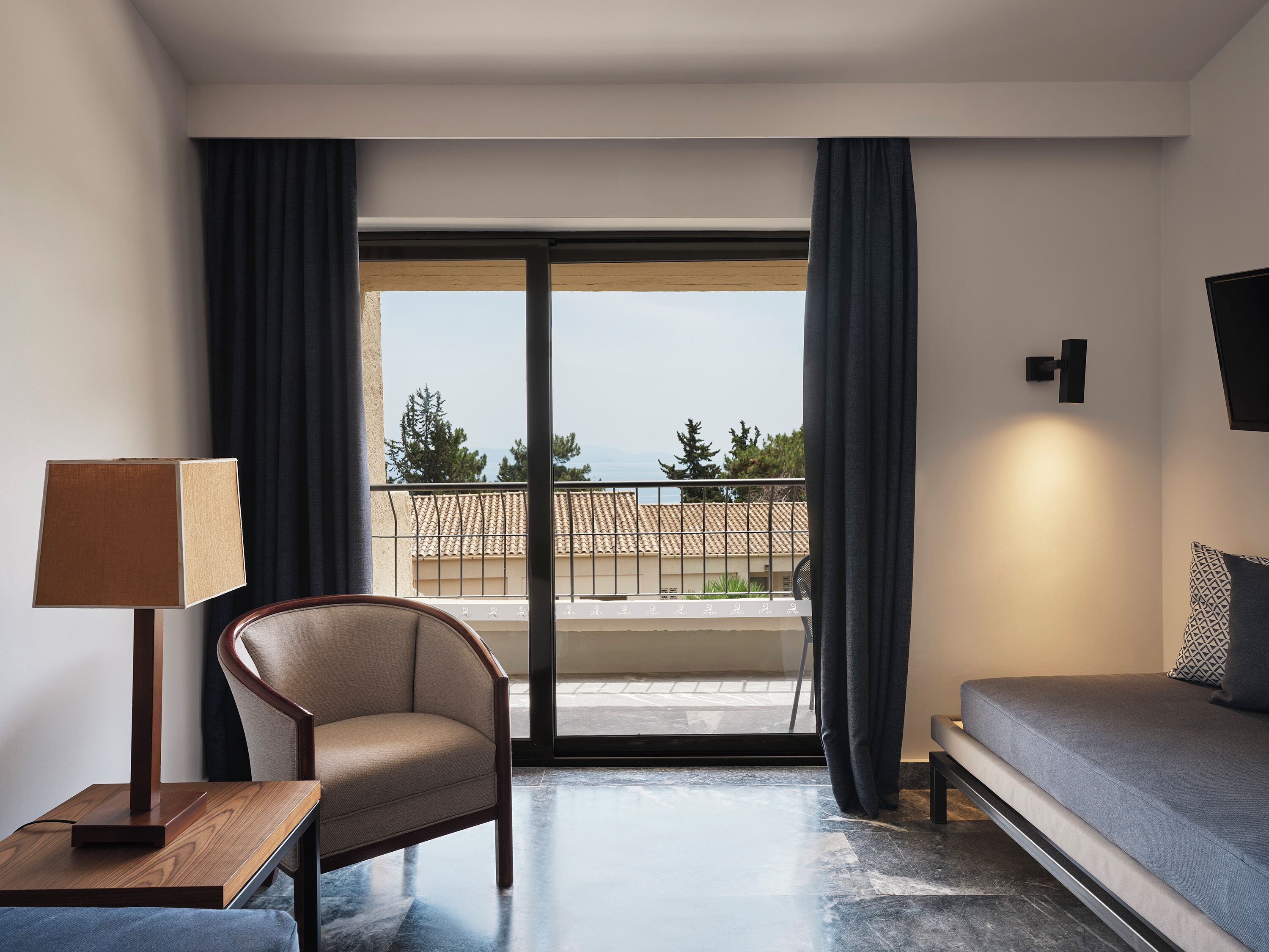 Een hotelkamer met een bank, een stoel, een lamp en uitzicht op zee door een groot raam.