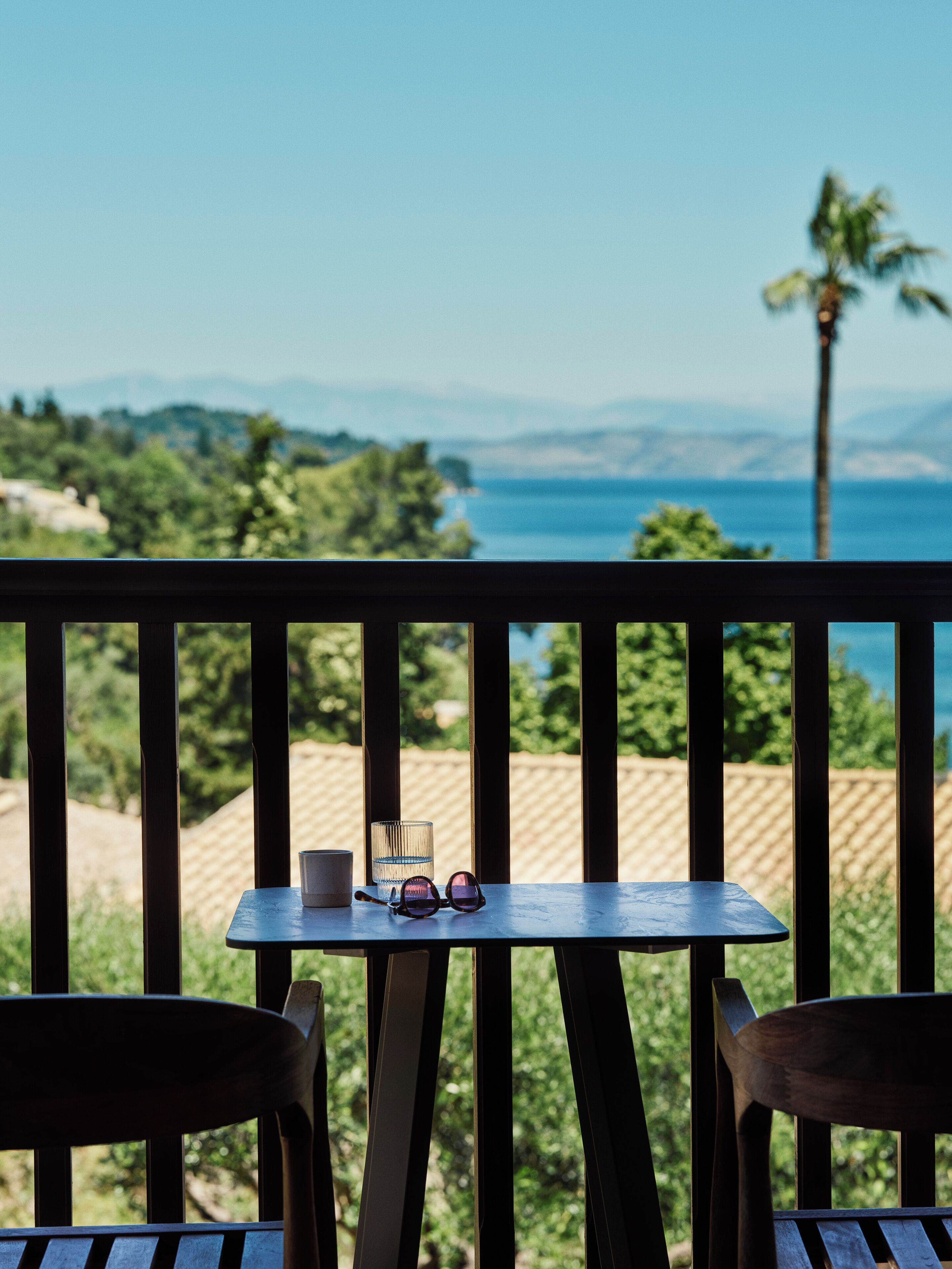 A balcony with a table and chairs, overlooking a scenic view of the sea and mountains.