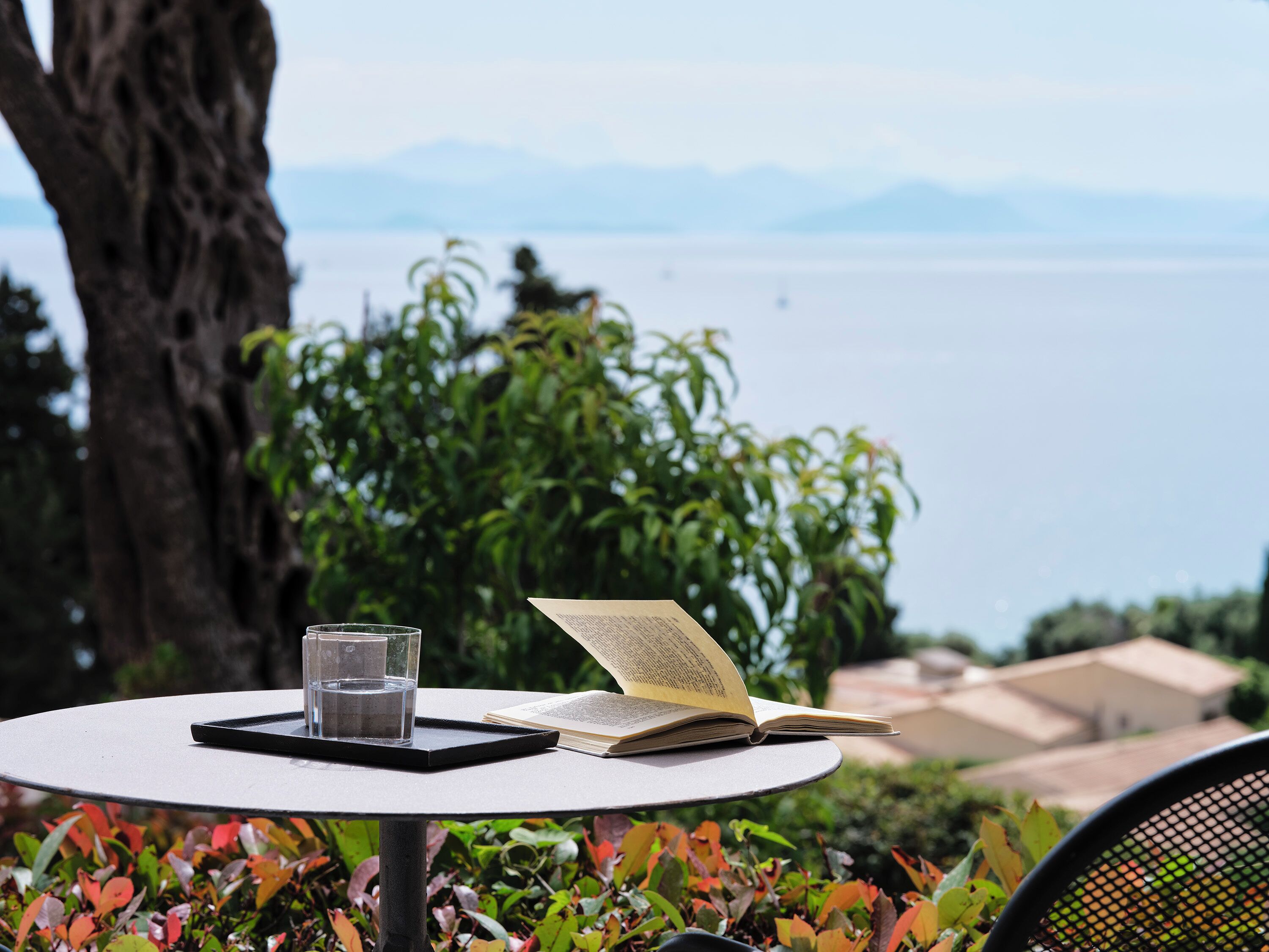 A table with a book and a glass of water, set against a backdrop of greenery and a view of the sea.