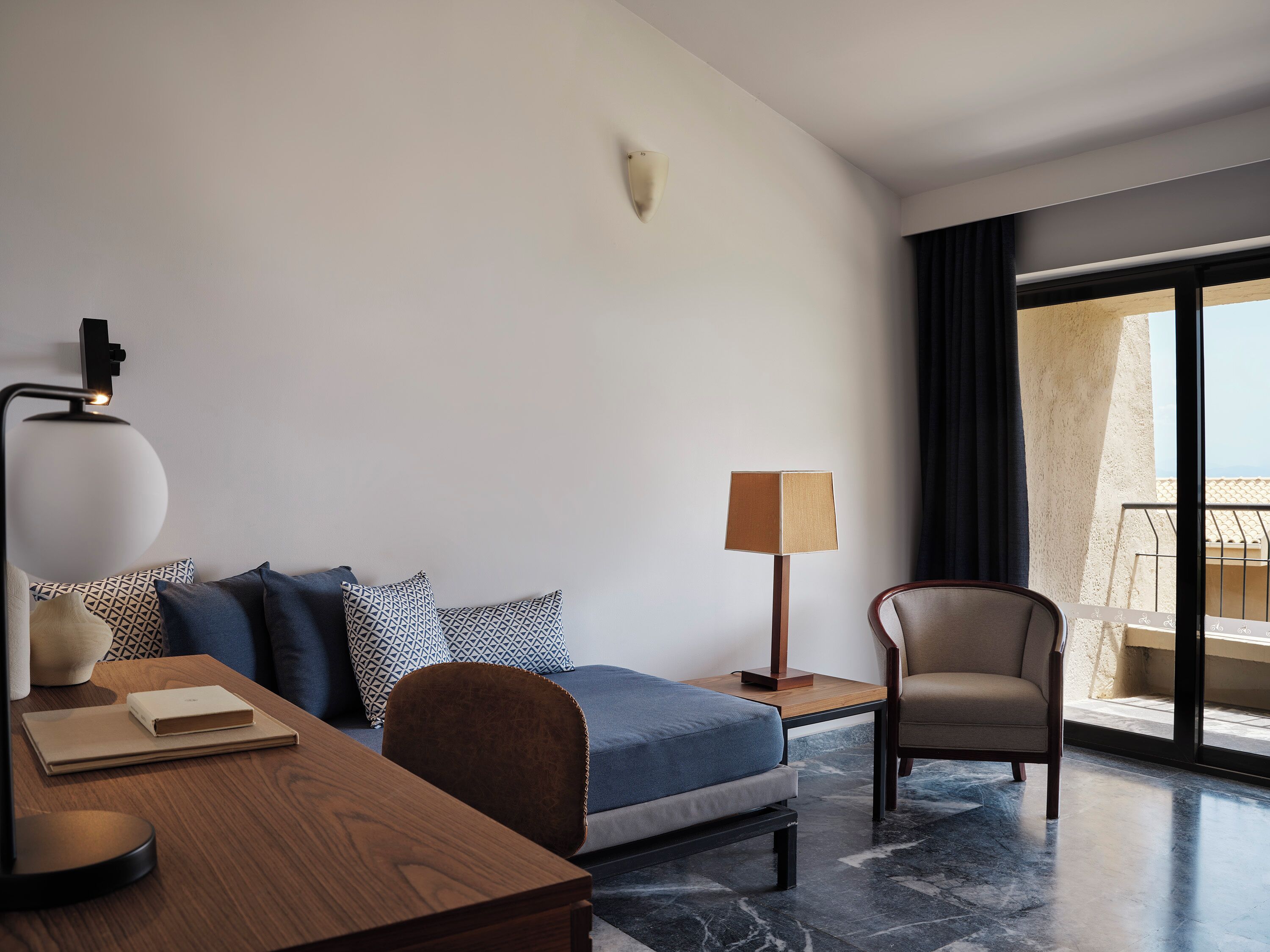 A modern hotel room with a bed, a desk, a chair, a lamp, and a view of the outside.