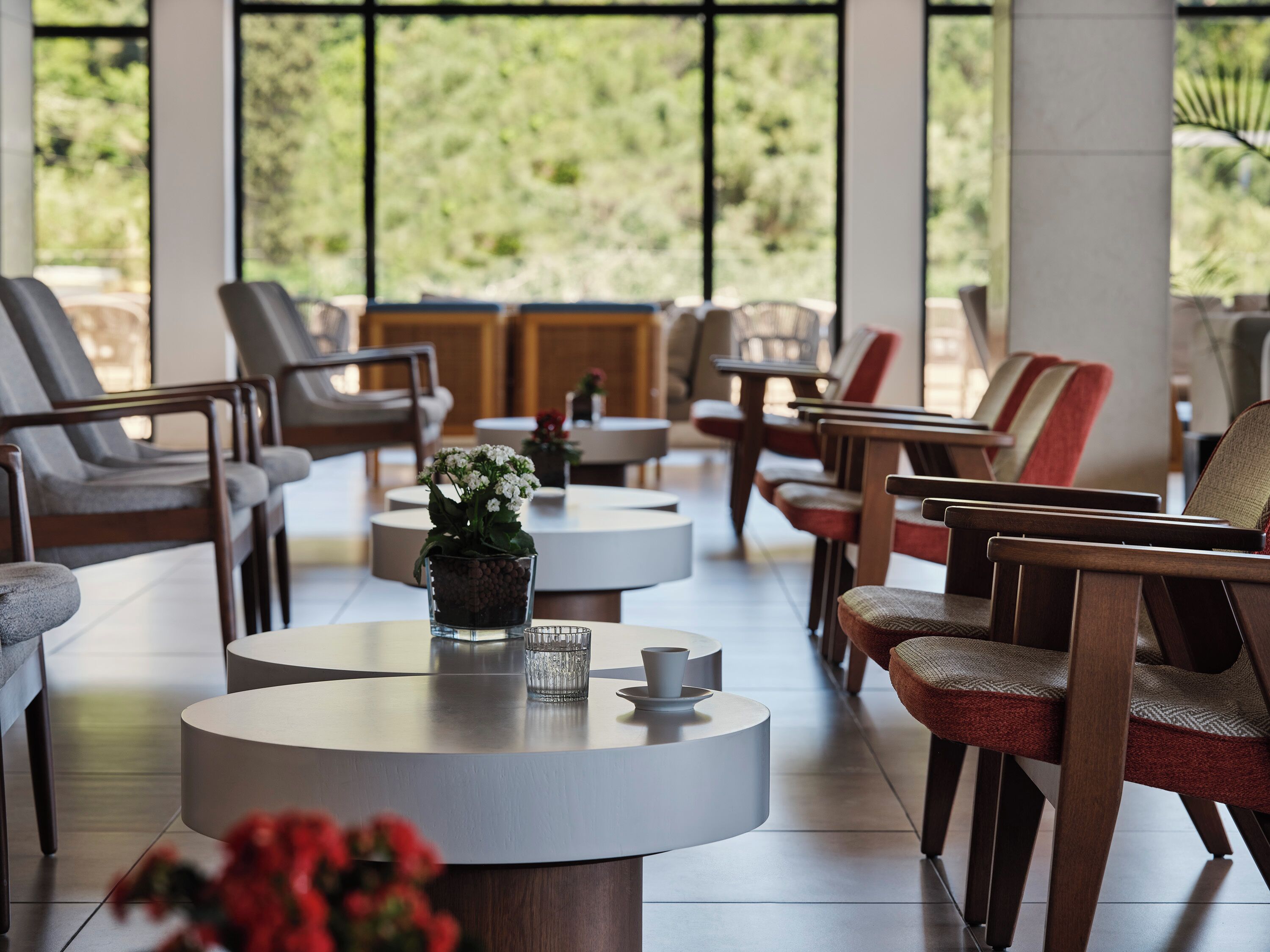 A modern lounge area with round tables, comfortable seating, and a view of greenery through large windows.