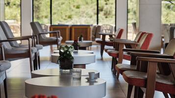 A modern lounge area with round tables, comfortable seating, and a view of greenery through large windows.