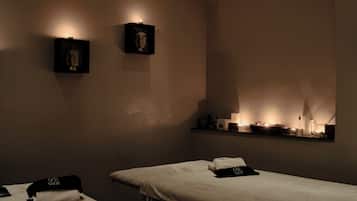 Ein Spa-Raum mit zwei Massageliegen, jeweils mit Handtuch und schwarzem Stirnband. An den Wänden sind Leuchten angebracht, und ein Regal beherbergt Kerzen und kleine Statuen.