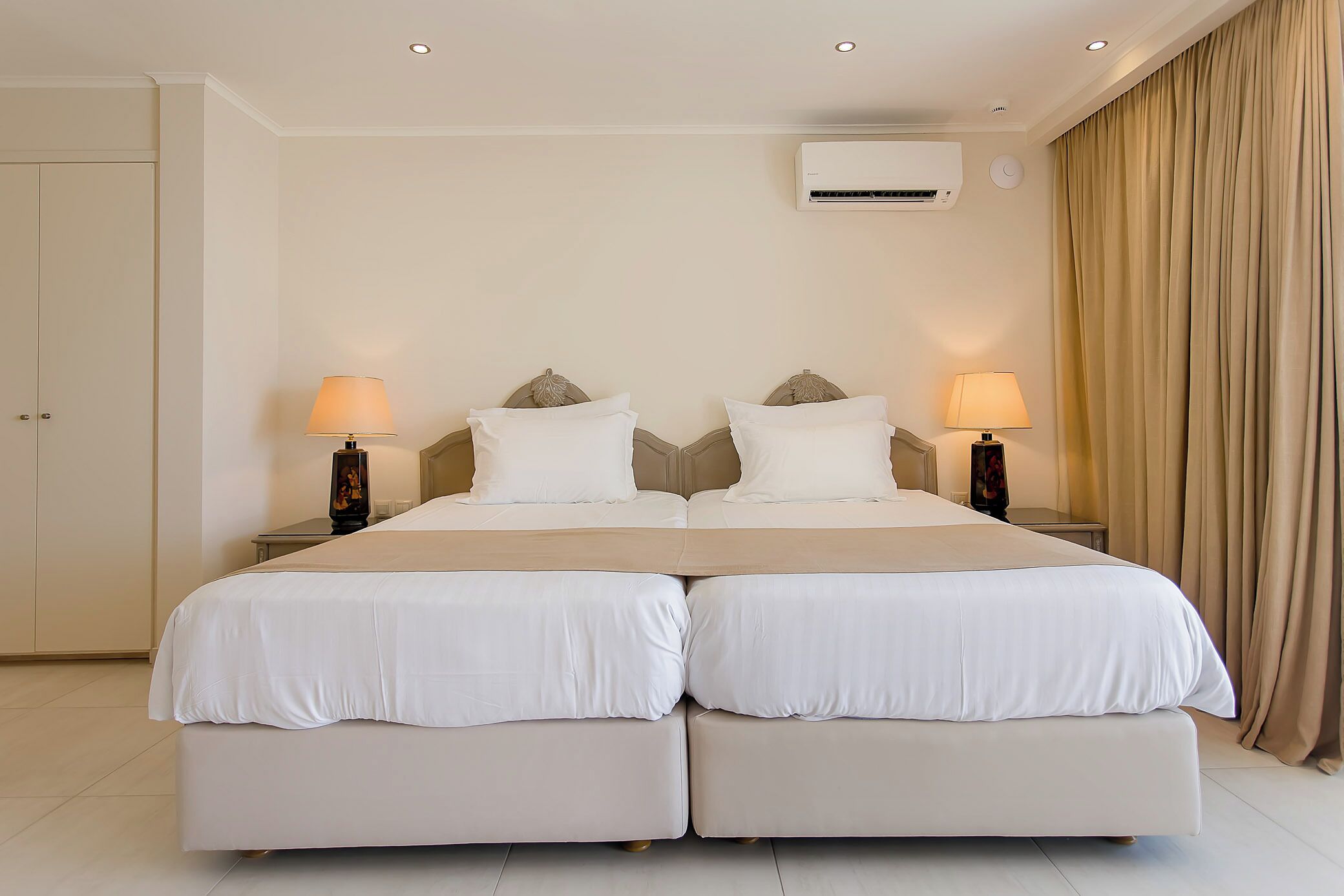 Una cama doble con ropa de cama blanca, dos mesitas de noche con lámparas, un aire acondicionado de pared y una puerta a la izquierda.