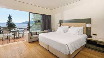 Habitación de hotel con una cama grande, un sofá, una mesita y un balcón con vistas al mar y las montañas.