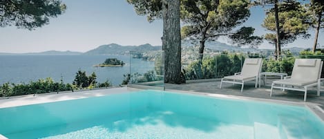 Une piscine d’un bleu éclatant, bordée de chaises longues blanches, et offrant une vue sur la mer et les montagnes.