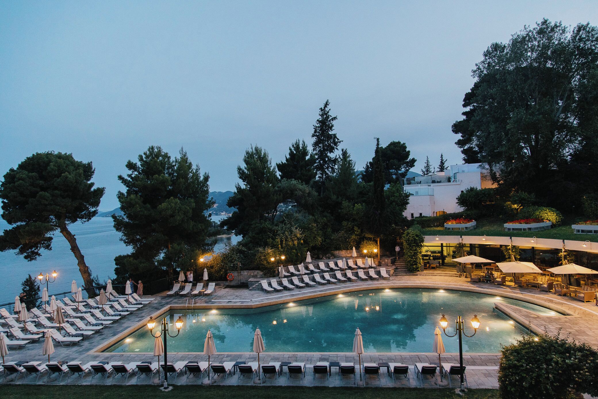 A poolside area with lounge chairs and umbrellas, surrounded by trees and overlooking a body of water.