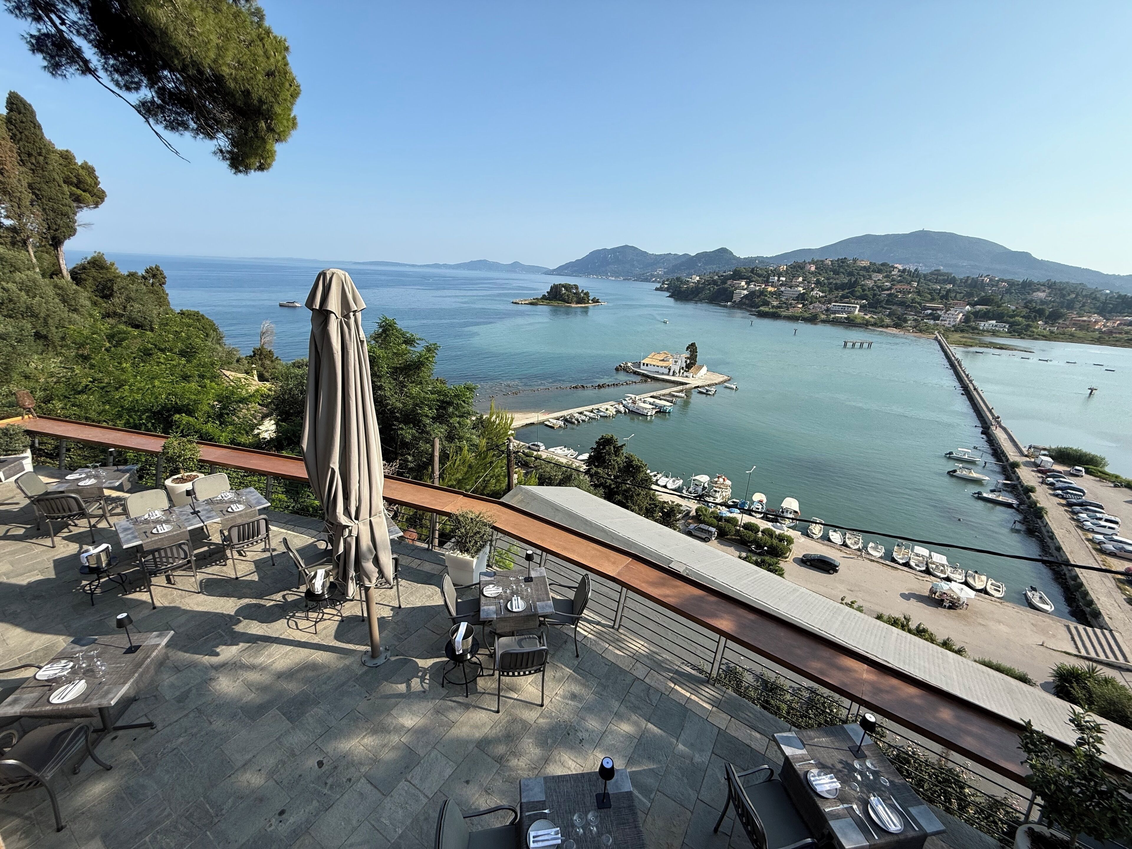 An outdoor terrace with tables and chairs, overlooking a marina with boats and a coastal town.