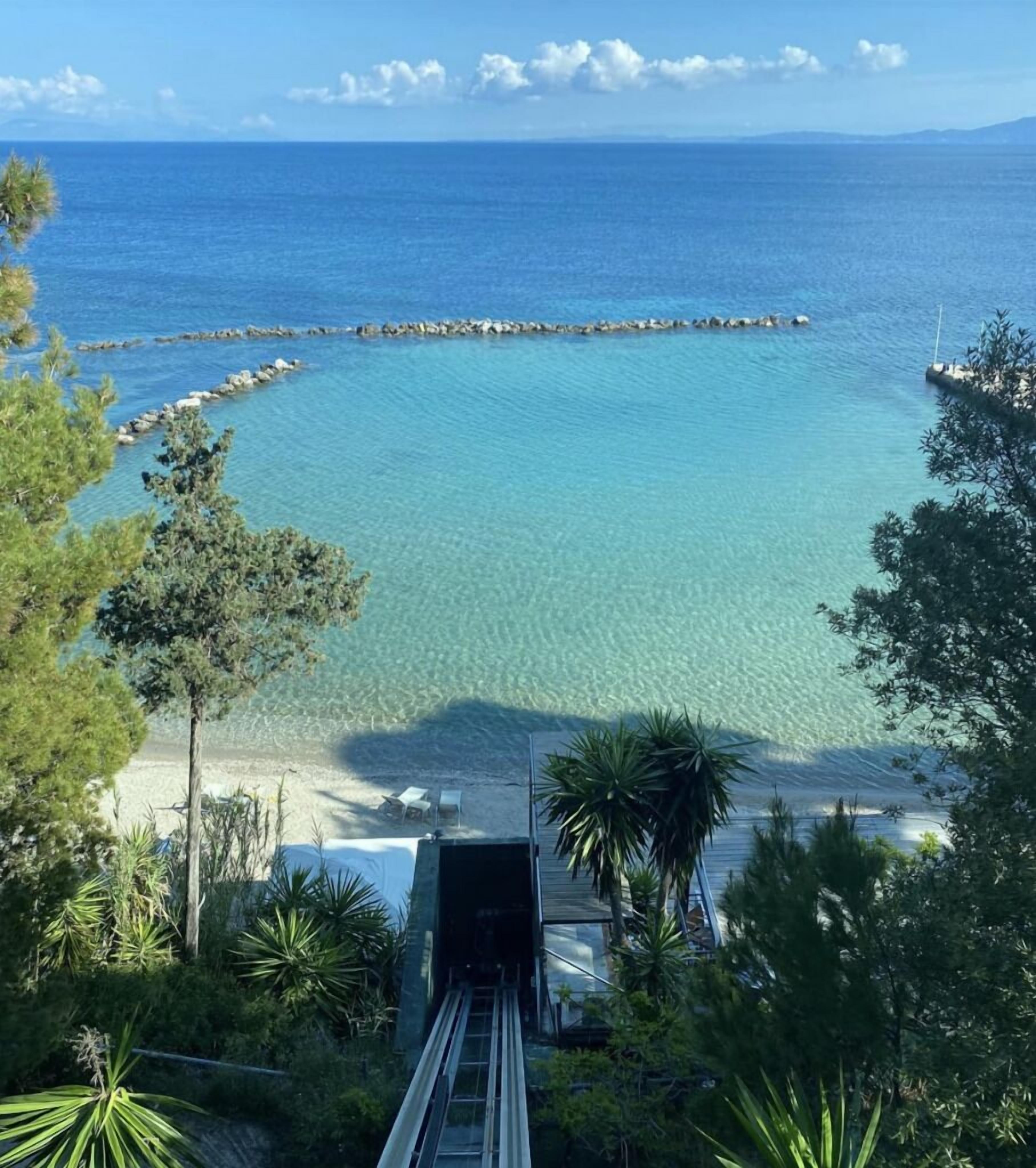 Vista costera con mar azul cristalino, un rompeolas rocoso y un camino que lleva a una zona de playa con sillas y sombrillas.