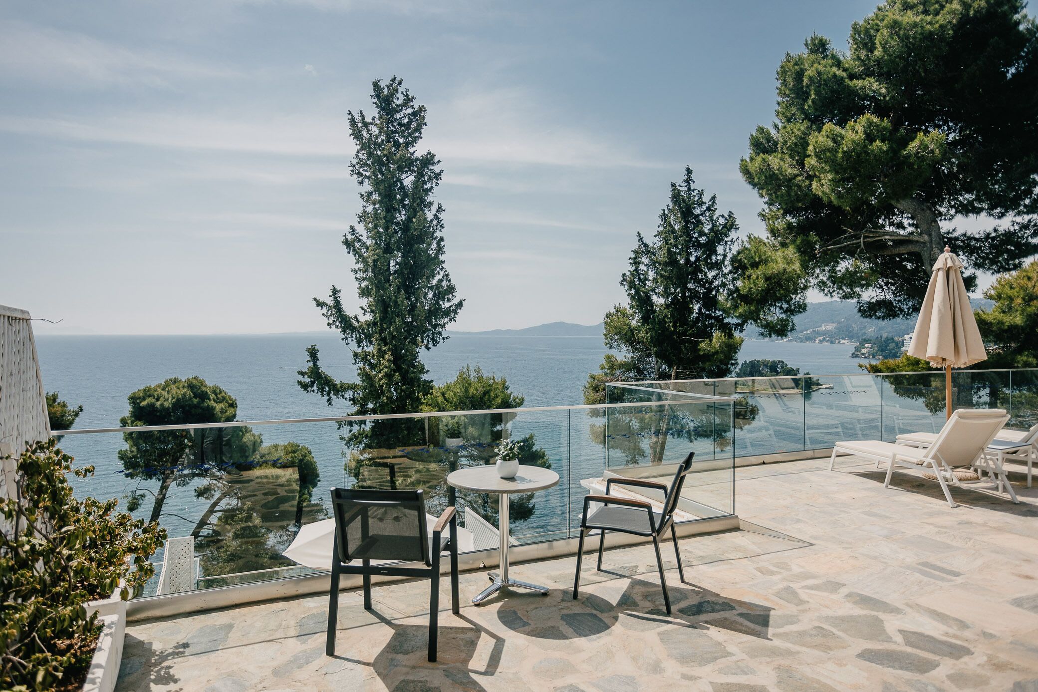 Une terrasse dotée d’une balustrade en verre donnant sur une étendue d’eau, aménagée avec des chaises et une table, et entourée d’arbres.