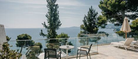 Une terrasse dotée d’une balustrade en verre donnant sur une étendue d’eau, aménagée avec des chaises et une table, et entourée d’arbres.