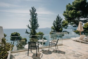 Una terraza con barandilla de cristal que da a un cuerpo de agua, amueblada con sillas y una mesa, y rodeada de árboles.