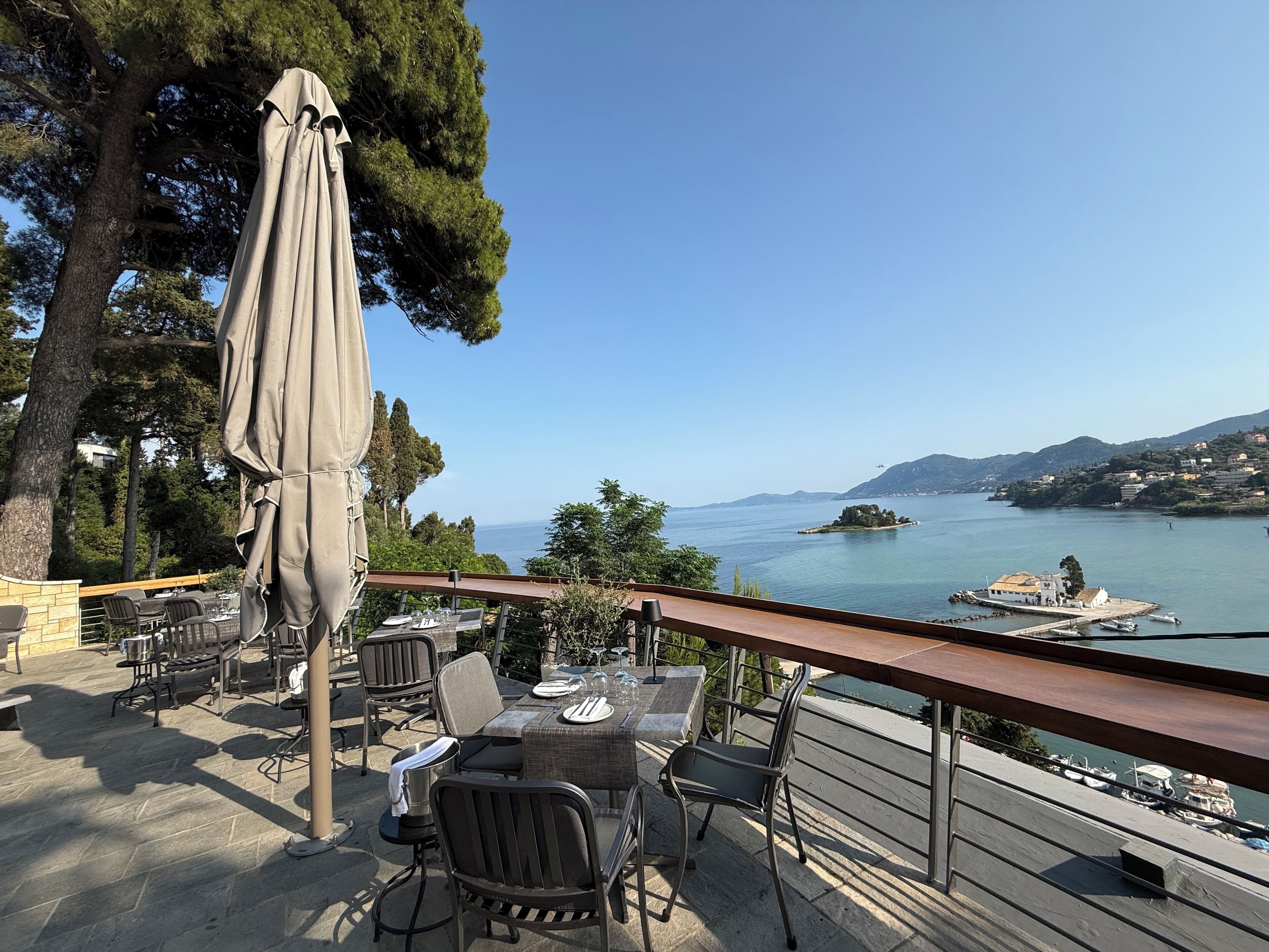 An outdoor dining area with tables and chairs, overlooking a body of water with boats and a distant island.