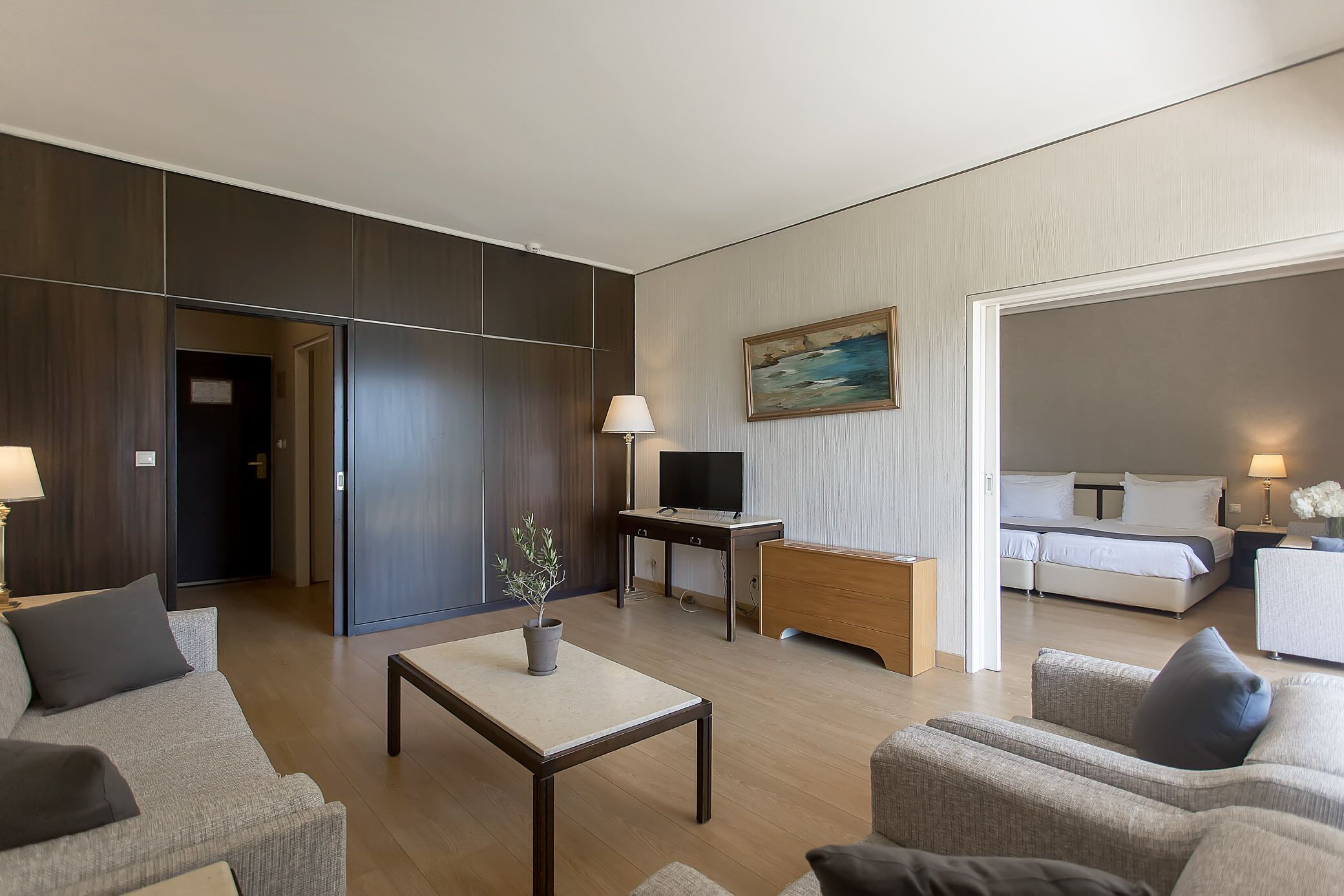 A modern hotel room with a sofa, armchair, coffee table, TV, bedside table, lamp, and a bed with a nightstand.