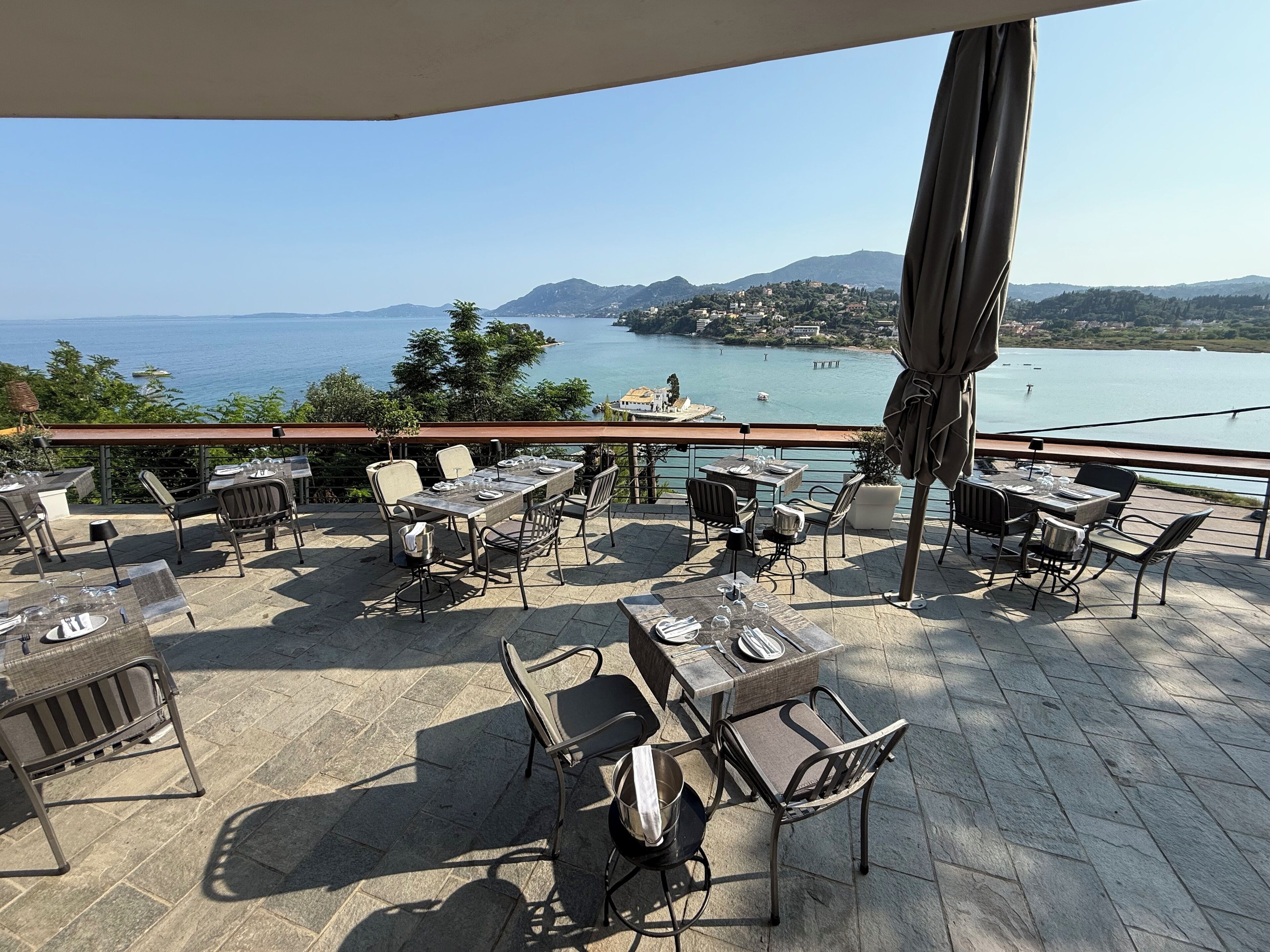 An outdoor terrace with tables and chairs, overlooking a body of water and distant hills.
