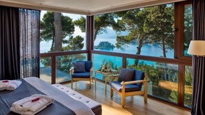 Habitación de hotel con un ventanal grande que ofrece vistas al mar y a árboles. Hay una cama con toallas y una mesita con un arreglo floral.