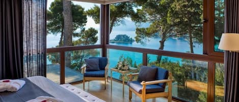 A hotel room with a large window offering a view of the sea and trees. There is a bed with towels and a small table with a flower arrangement.