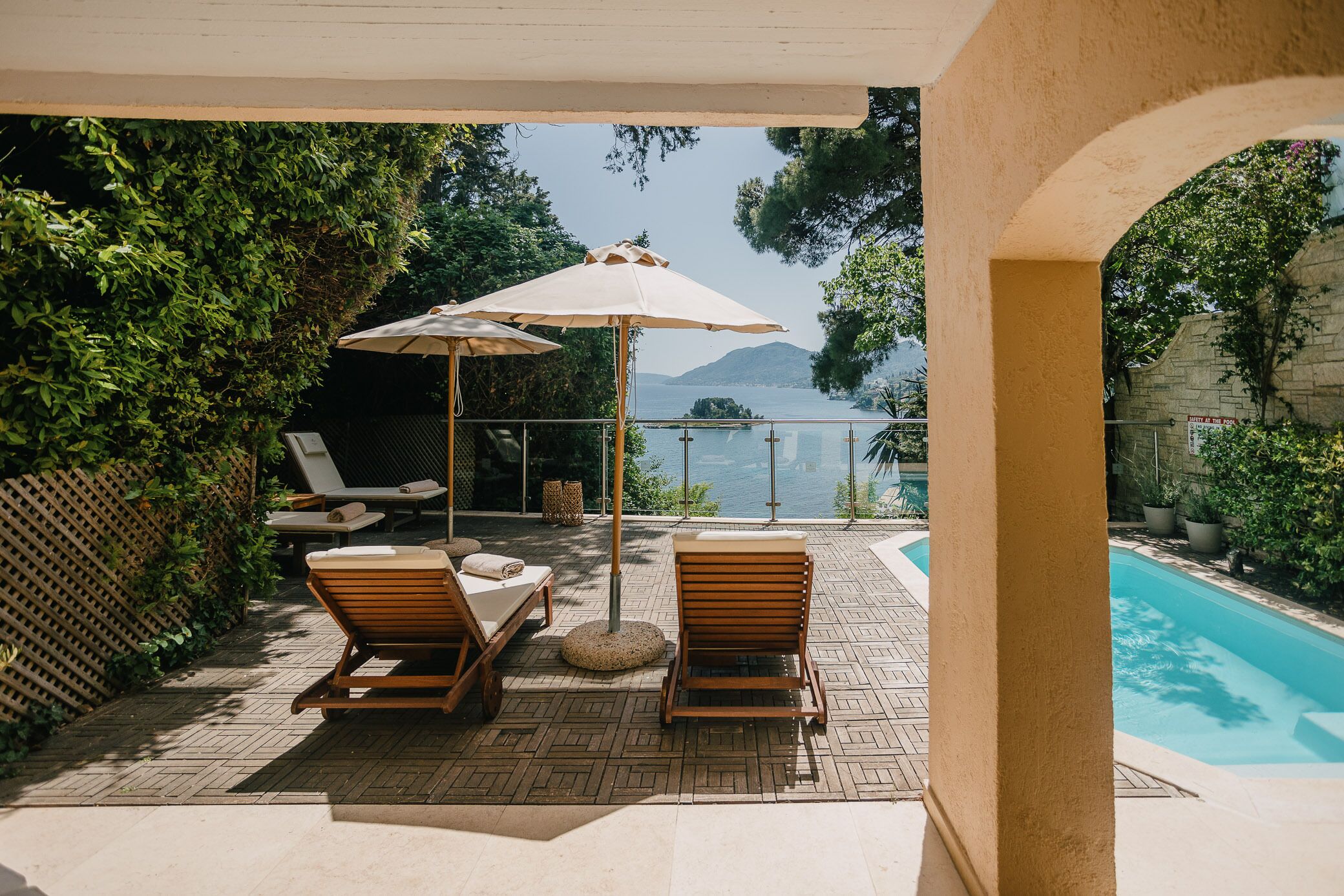 Un espace aménagé au bord de la piscine, avec des chaises longues et un parasol, offrant une vue sur une étendue d’eau et la verdure environnante.