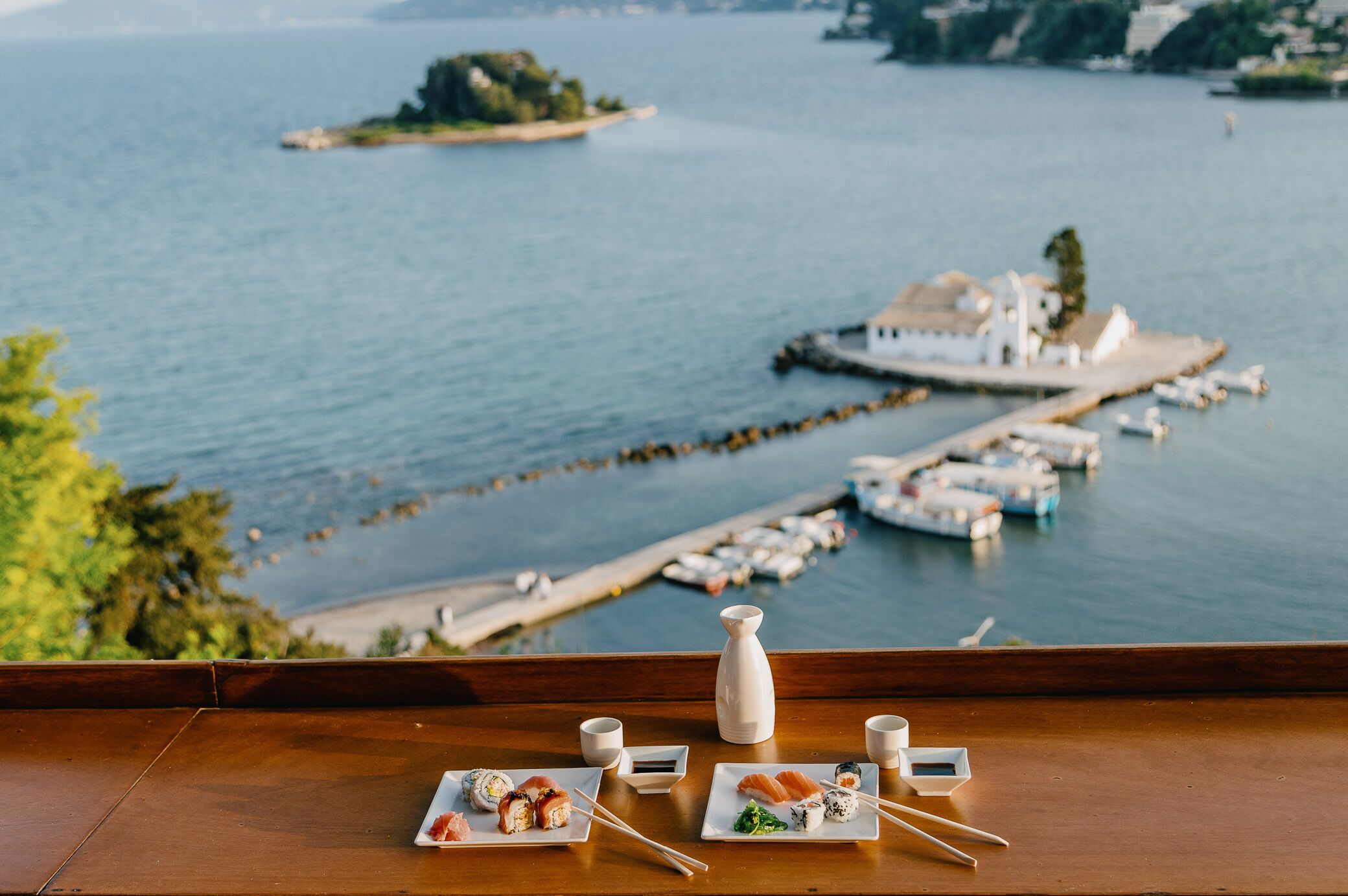 A dining table with sushi plates and a vase, overlooking a marina with boats and a distant island.