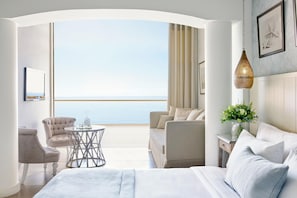 A modern hotel room with a balcony, a bed, a seating area, and a view of the sea.