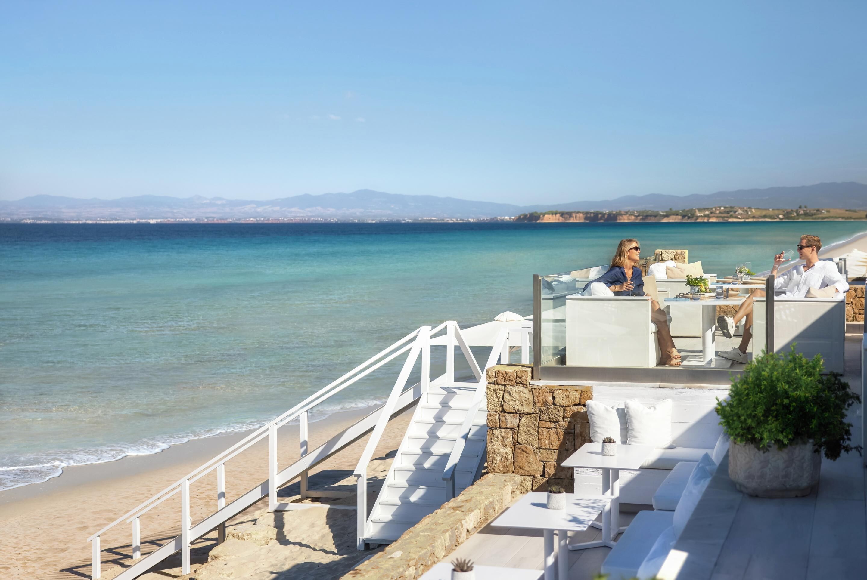Eine Strandterrasse mit Außenmöbeln, einer Steinmauer und Blick auf das Meer und ferne Berge.
