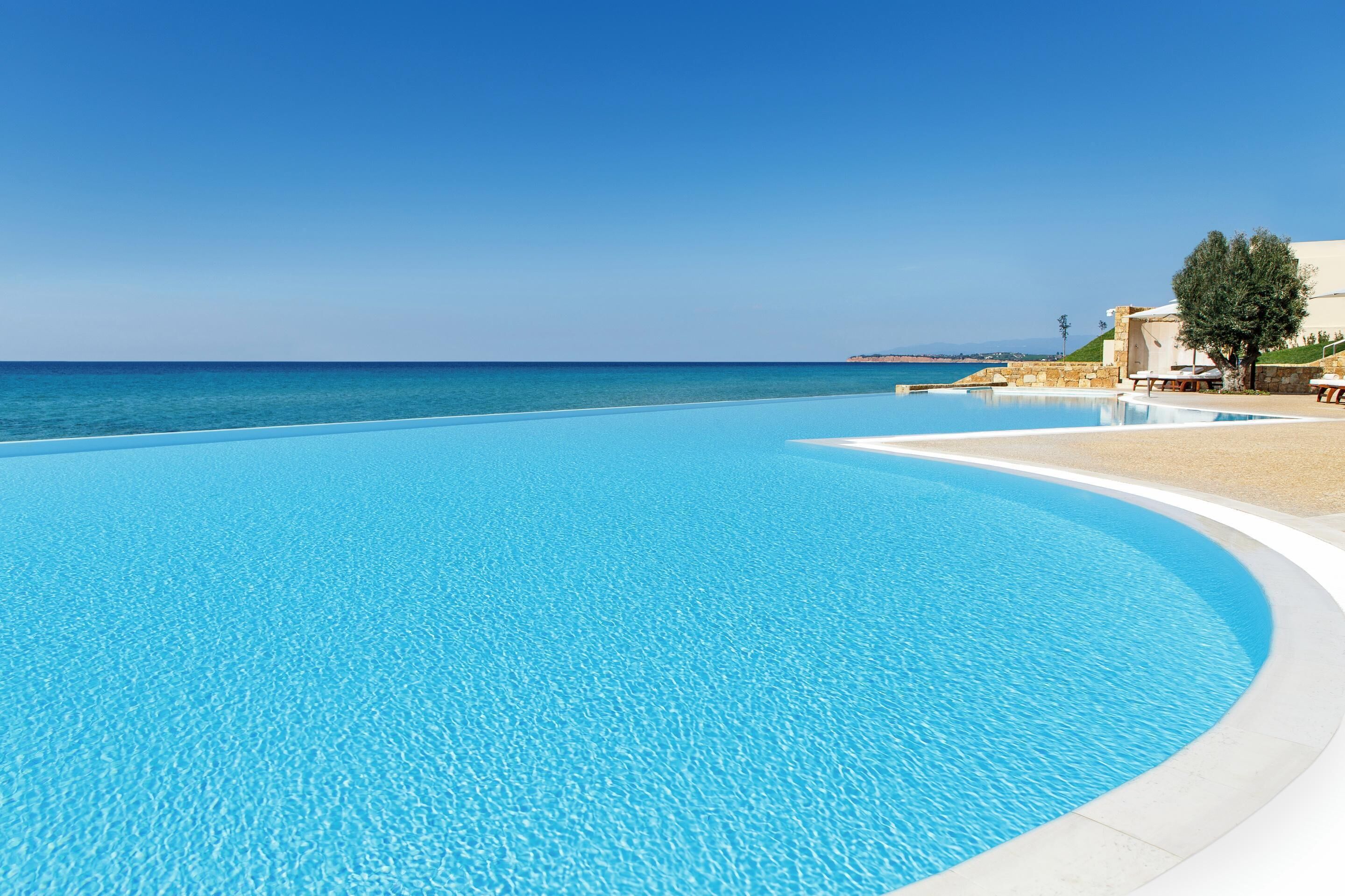 A clear blue pool with a curved edge, a sandy beach area, and a building with a white facade and a tree in the background.