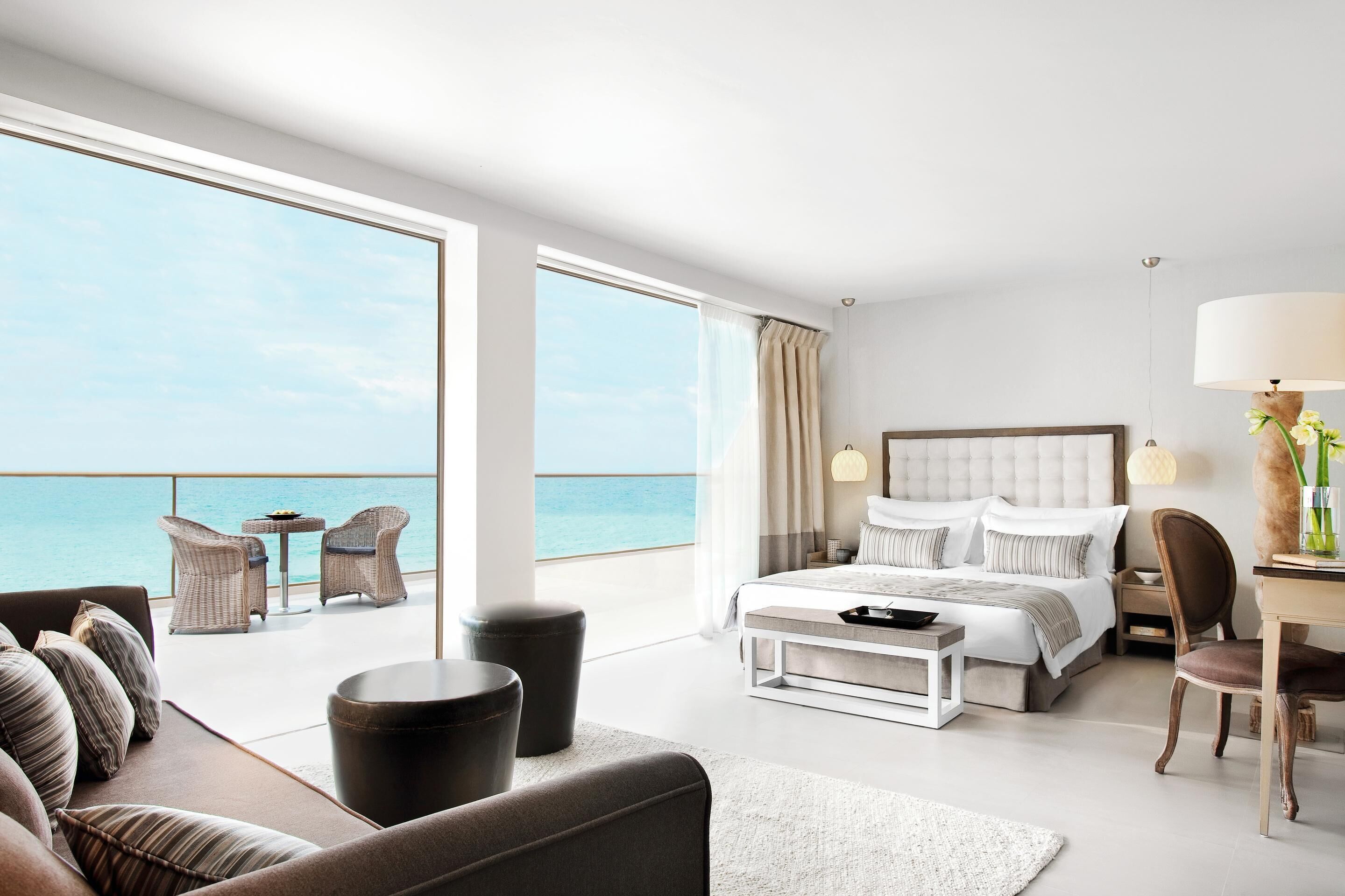 A modern hotel room with a large bed, a sofa, a dining area, and a balcony with a view of the sea.