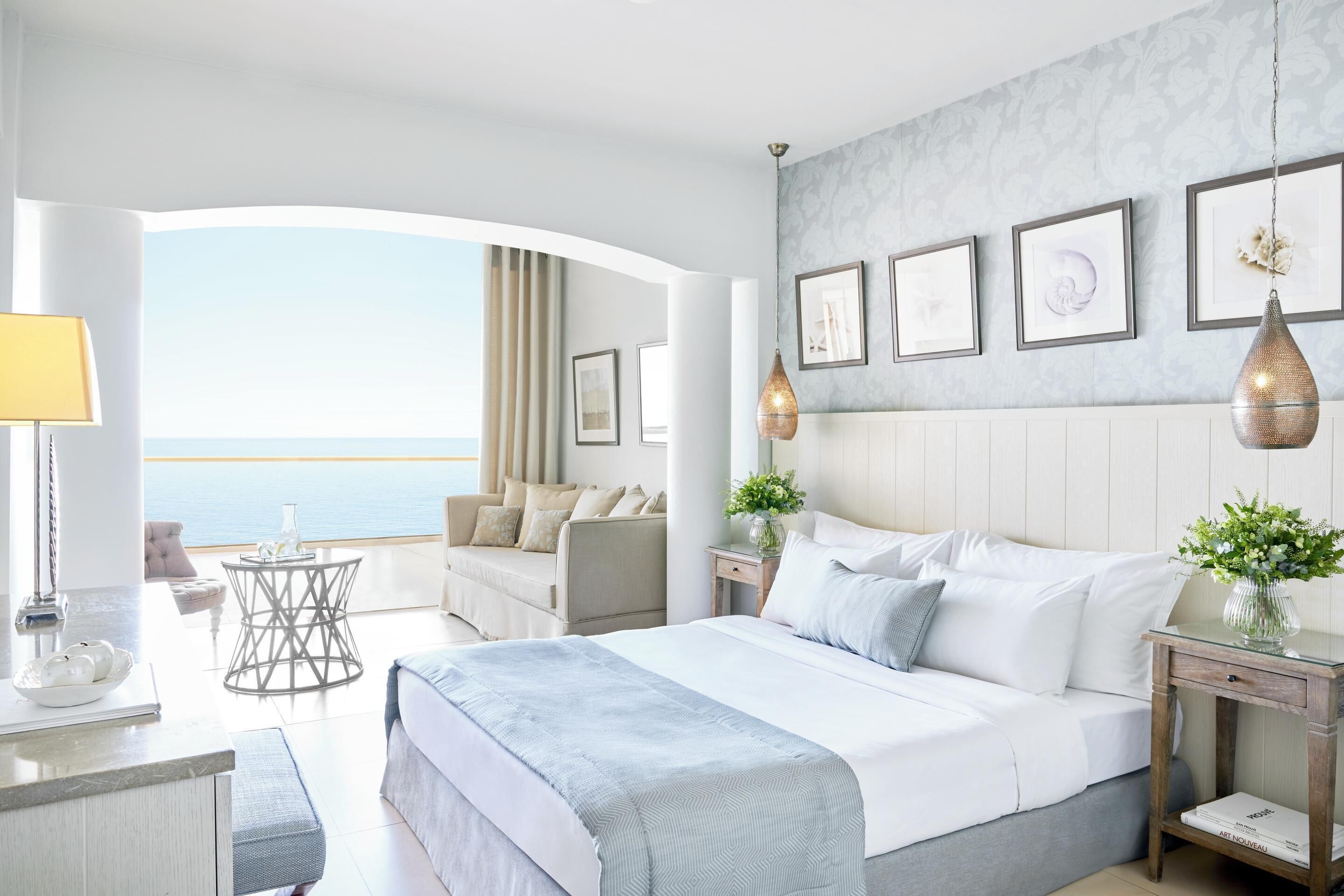 A modern hotel room with a large bed, a seating area with a view of the sea, and a balcony.