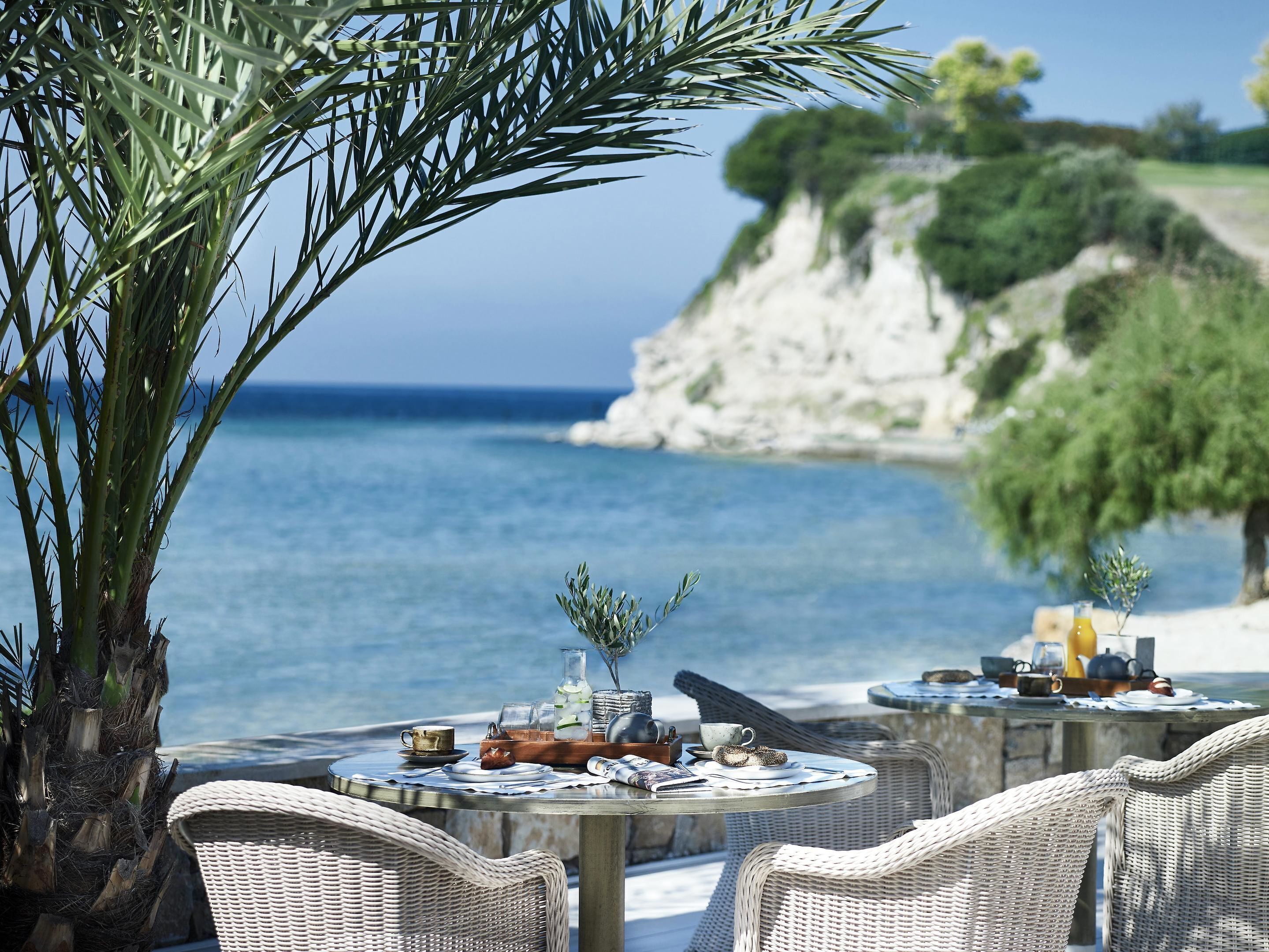 A seaside dining area with wicker chairs, a table set with a teapot, cups, and a bottle, overlooking the sea and a rocky cliff.