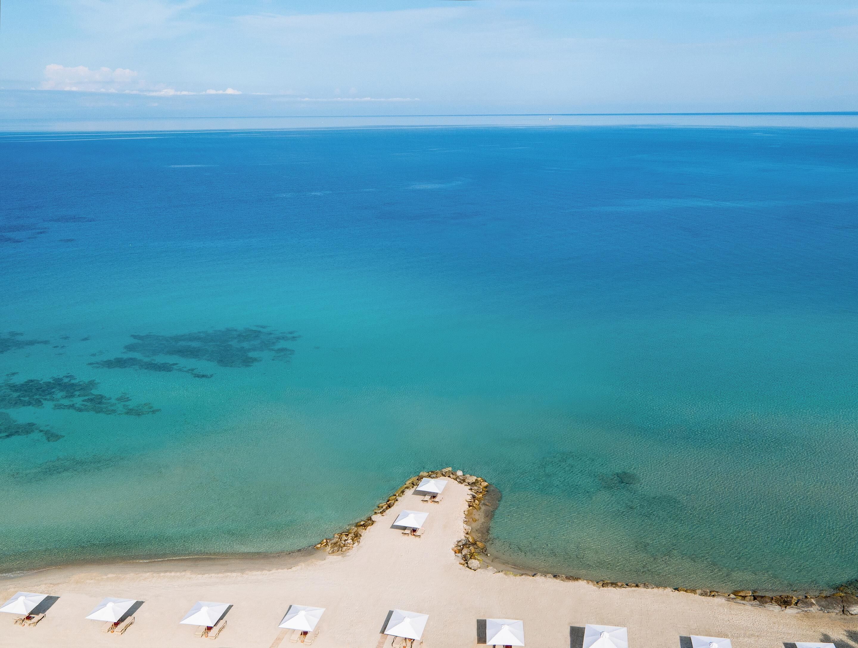 An aerial view of a beach with a row of white umbrellas and lounge chairs, a rocky breakwater, and clear blue water.