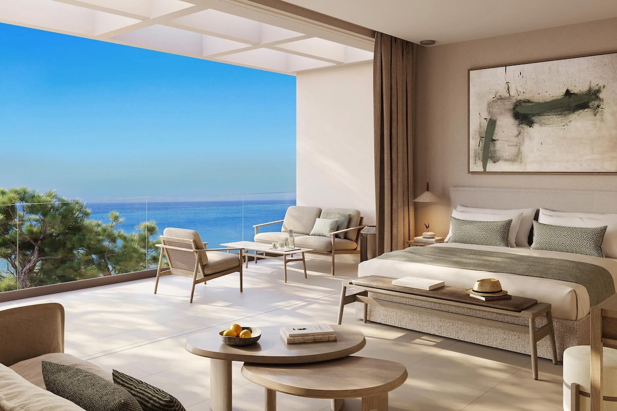 Quarto de hotel moderno com uma varanda grande, uma cama com várias almofadas, uma mesa de centro e vista para o mar.