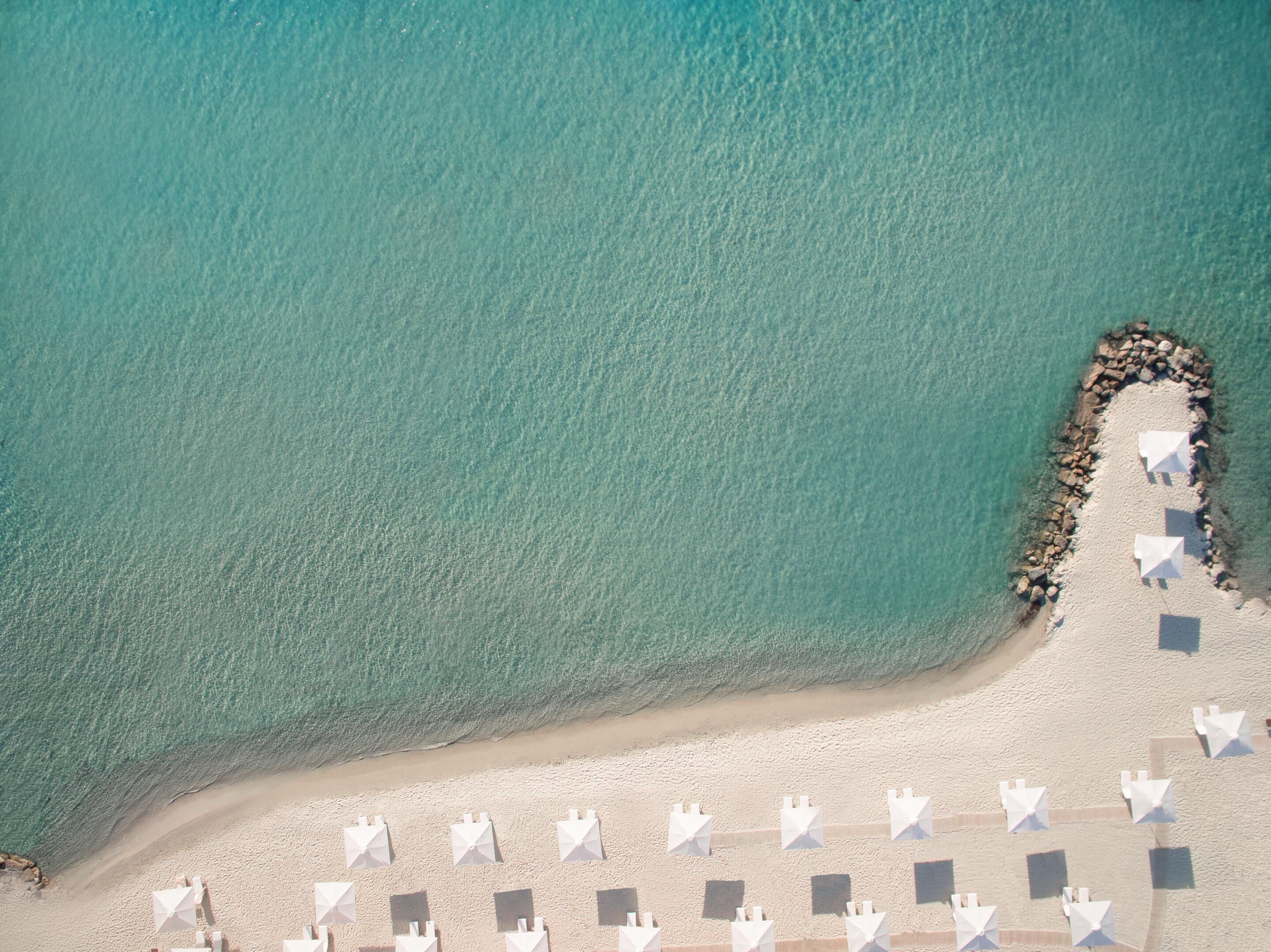 An aerial view of a beach with a row of white umbrellas and a rocky breakwater.