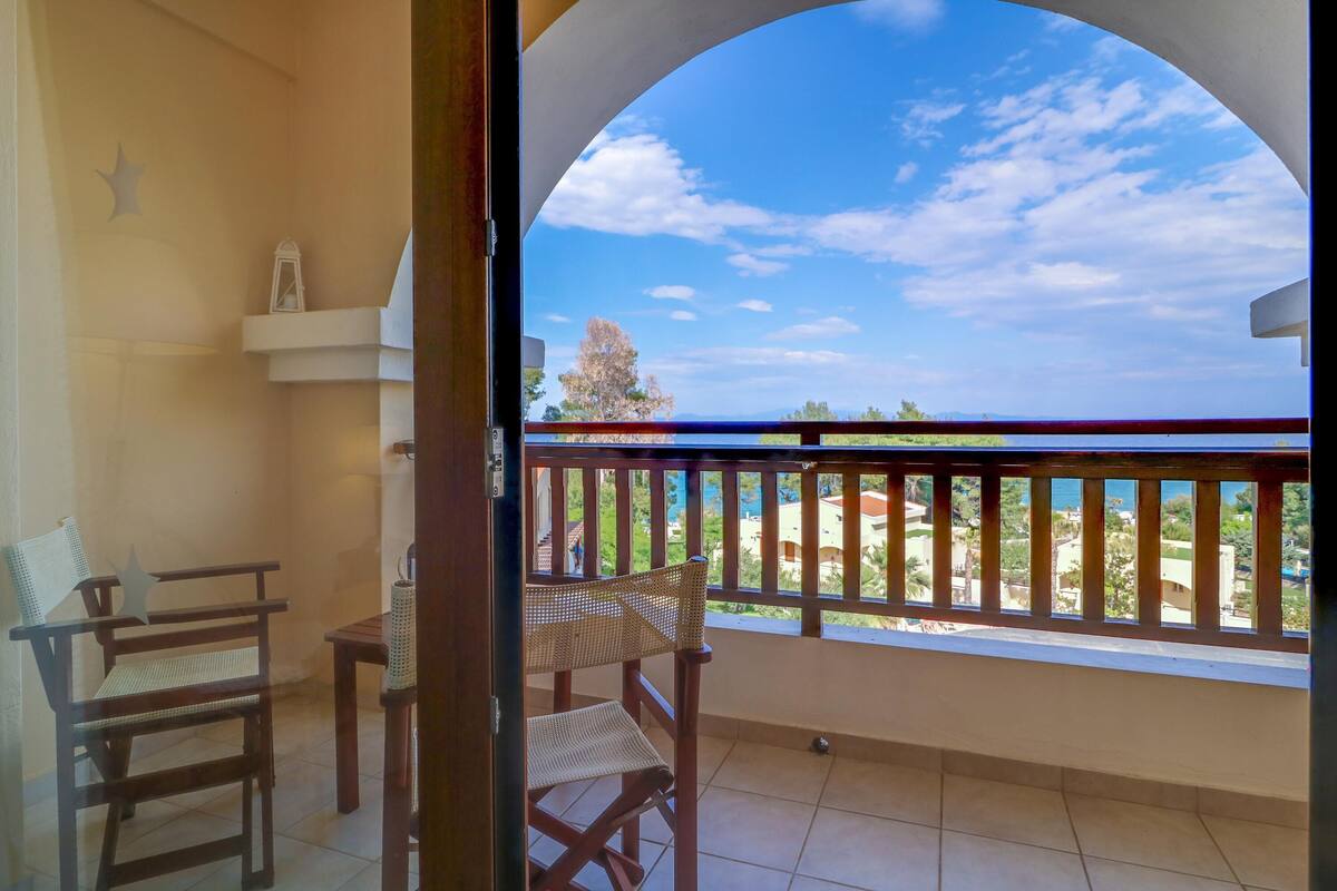 Un balcon avec vue sur la mer et un arbre. On y trouve deux chaises en bois et une petite table.